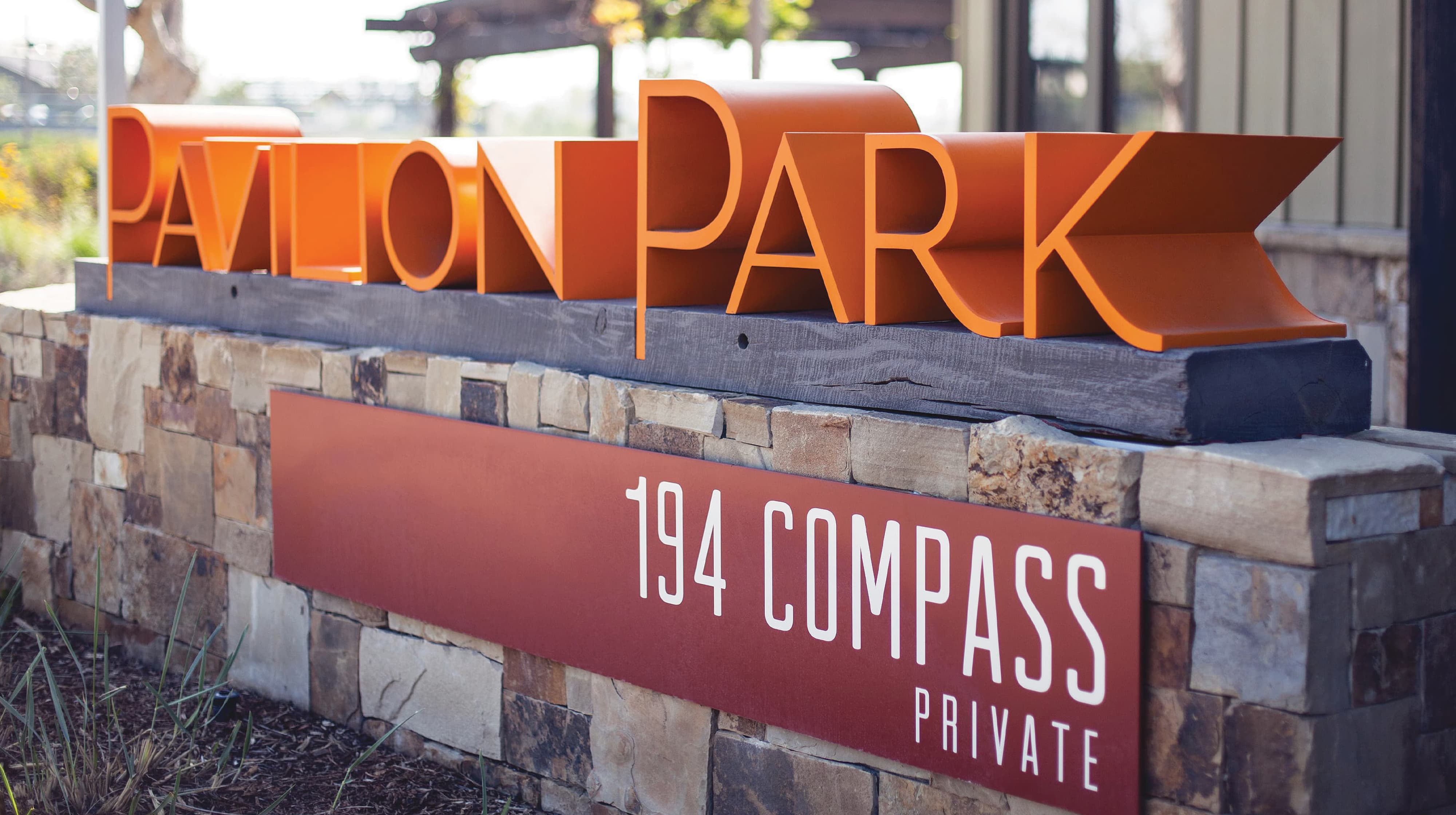 Dimensional orange 'Pavilion Park' sign with folded ribbon-style lettering on a stone wall, above a maroon '194 Compass Private' address plaque.