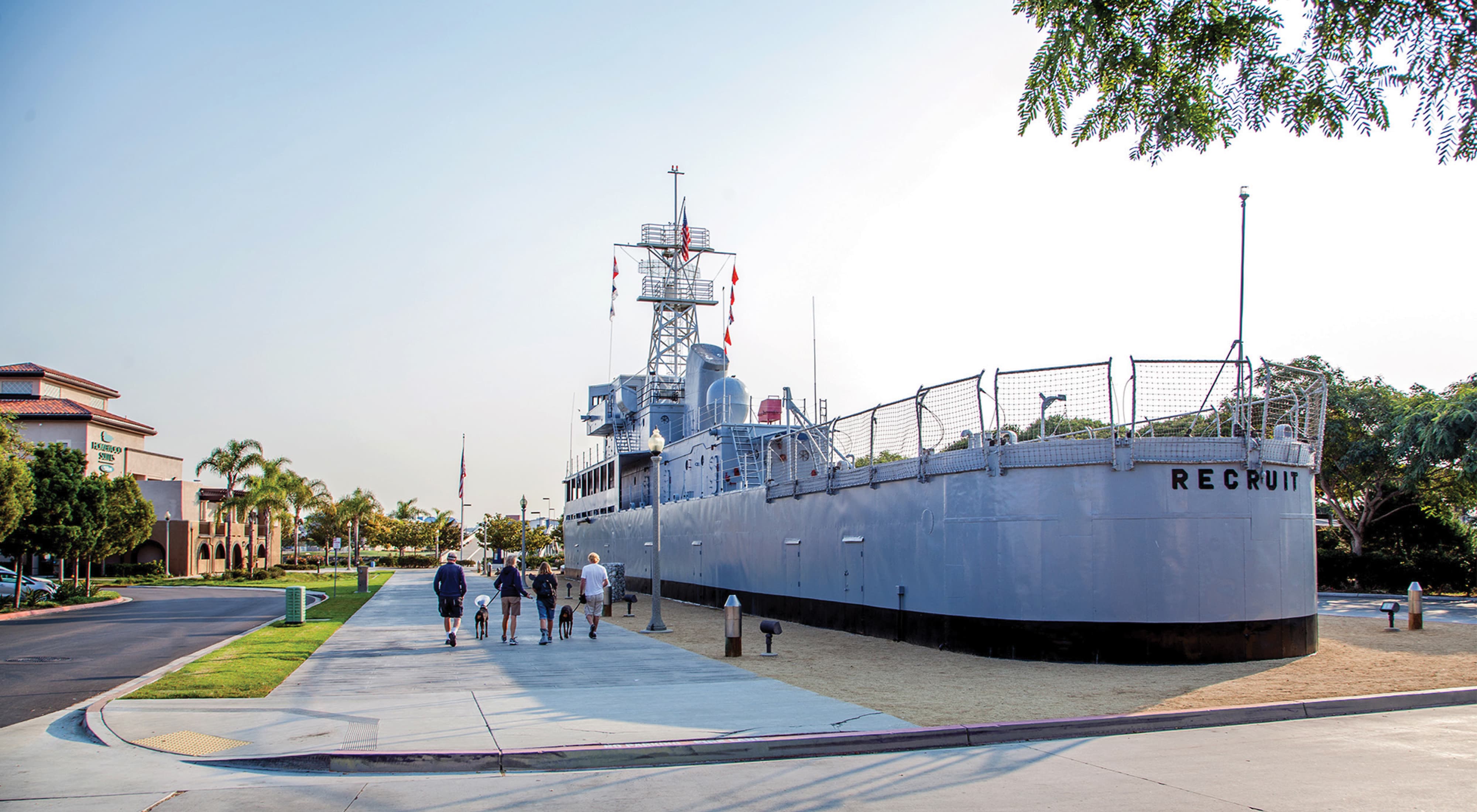 People walking dogs along a paved path beside the USS Recruit, a landlocked training ship hull painted gray with 'Recruit' lettering, palm trees and buildings in the background at golden hour.