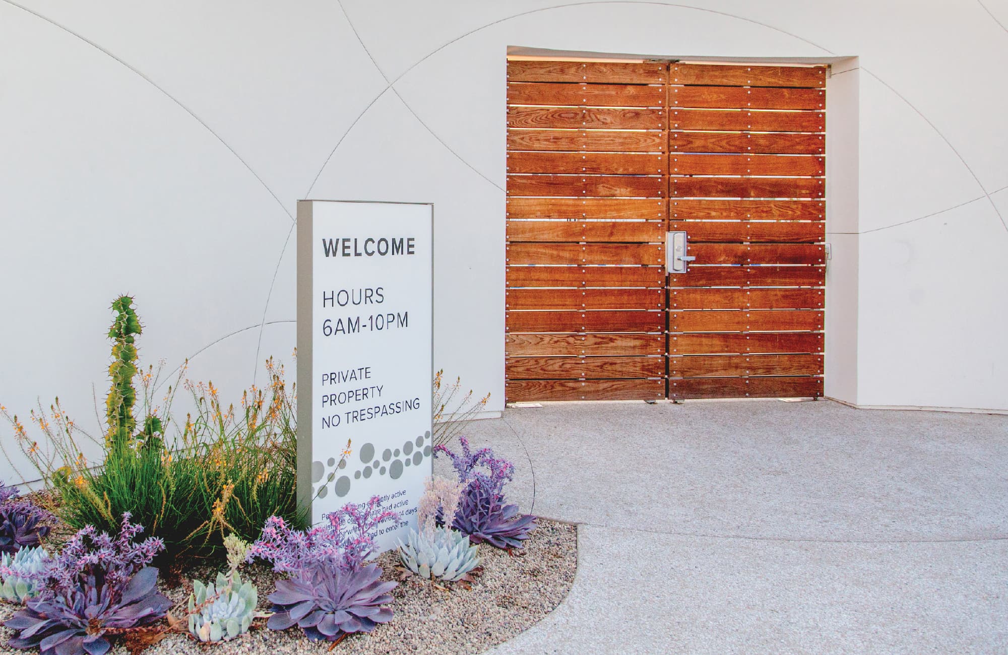 Entrance to a private community pool area with wood-plank double doors set into a curved white wall, beside a welcome sign listing hours and property rules, surrounded by cacti, succulents, and desert landscaping.