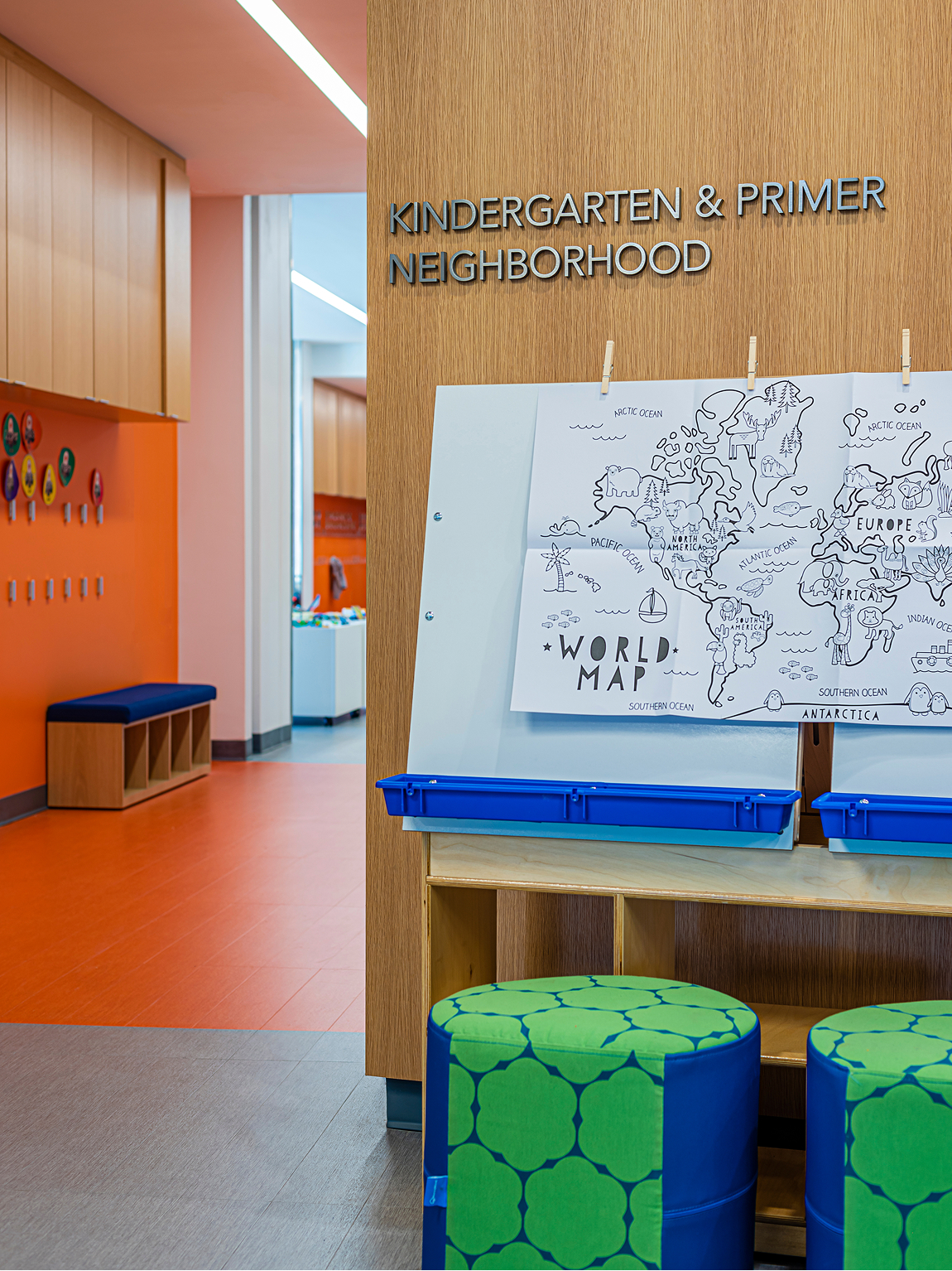 Wood wall reading 'Kindergarten & Primer Neighborhood' beside an illustrated world map easel with blue supply trays and green polka-dot ottomans, opening into a bright orange hallway with cubbies and a bench.