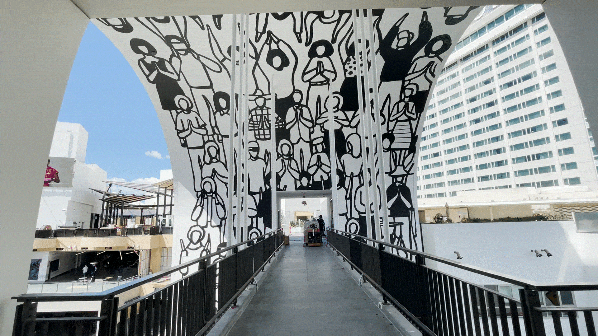 Immersive interior view looking down a pedestrian bridge corridor at Ovation Hollywood featuring a dramatic large-scale black and white graphic mural of stylized human figures, families, and community scenes wrapping across the curved overhead canopy and structural columns, creating an engaging public art installation along the open-air walkway with the Loews Hollywood Hotel tower visible in the background