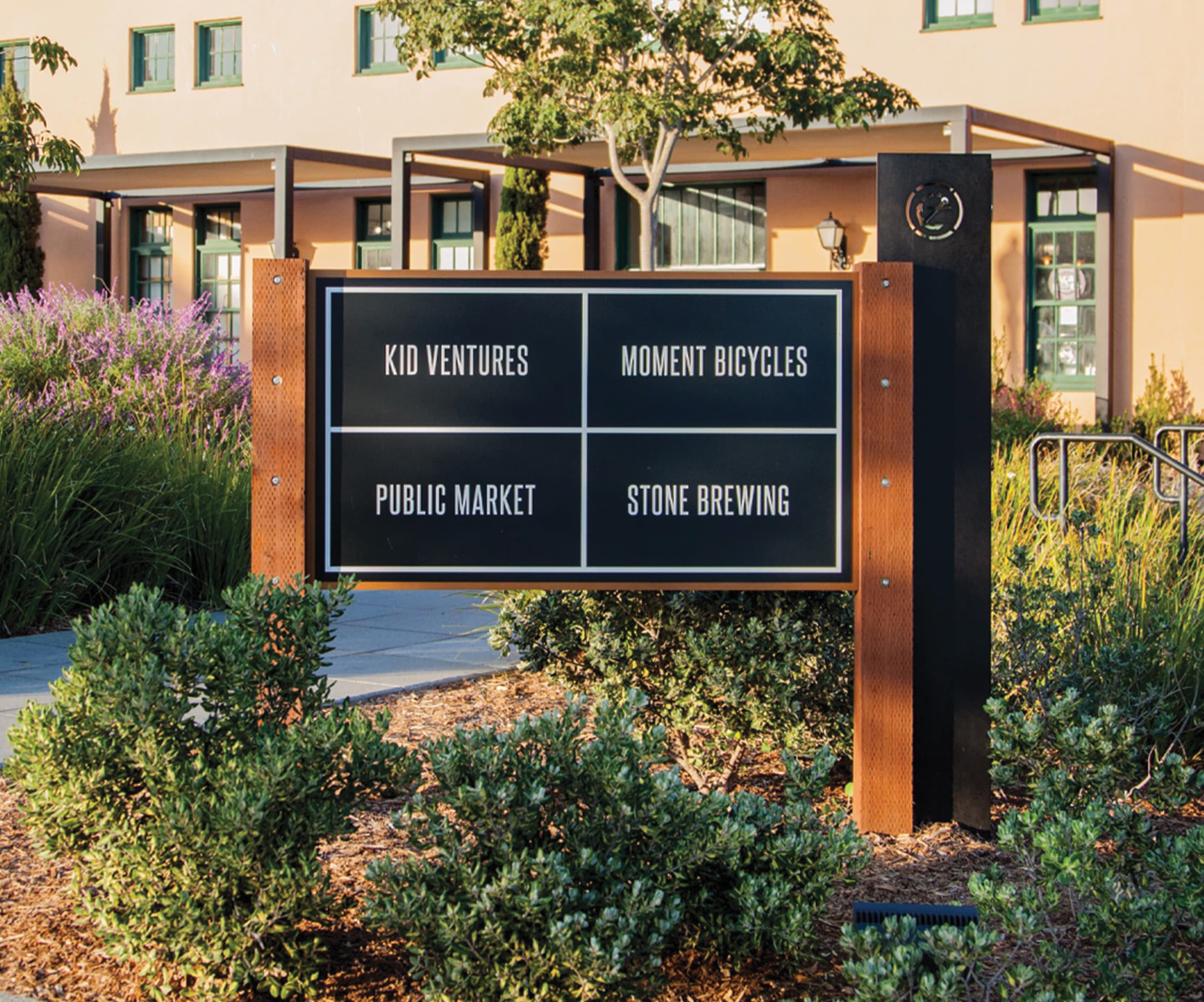 Four-panel directional sign with corten steel posts reading 'Kid Ventures, Moment Bicycles, Public Market, Stone Brewing,' set among flowering shrubs and grasses in front of a Spanish Colonial style building with green-trimmed windows.
