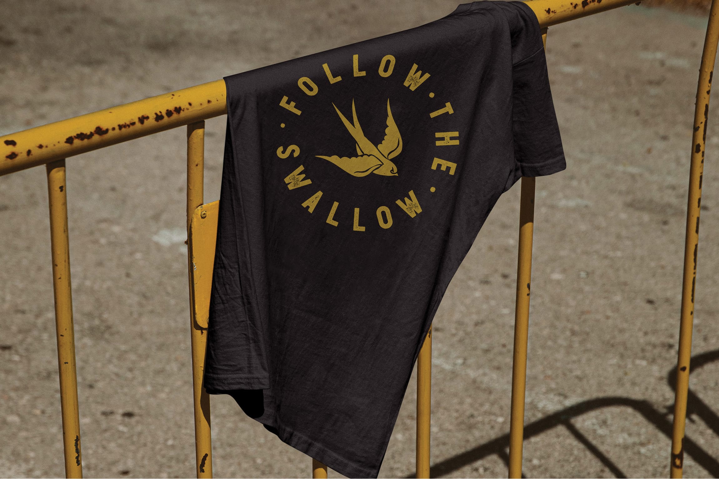 Black t-shirt with gold "Follow the Swallow" badge logo draped over a rusted yellow railing, brand identity design by RSM Design