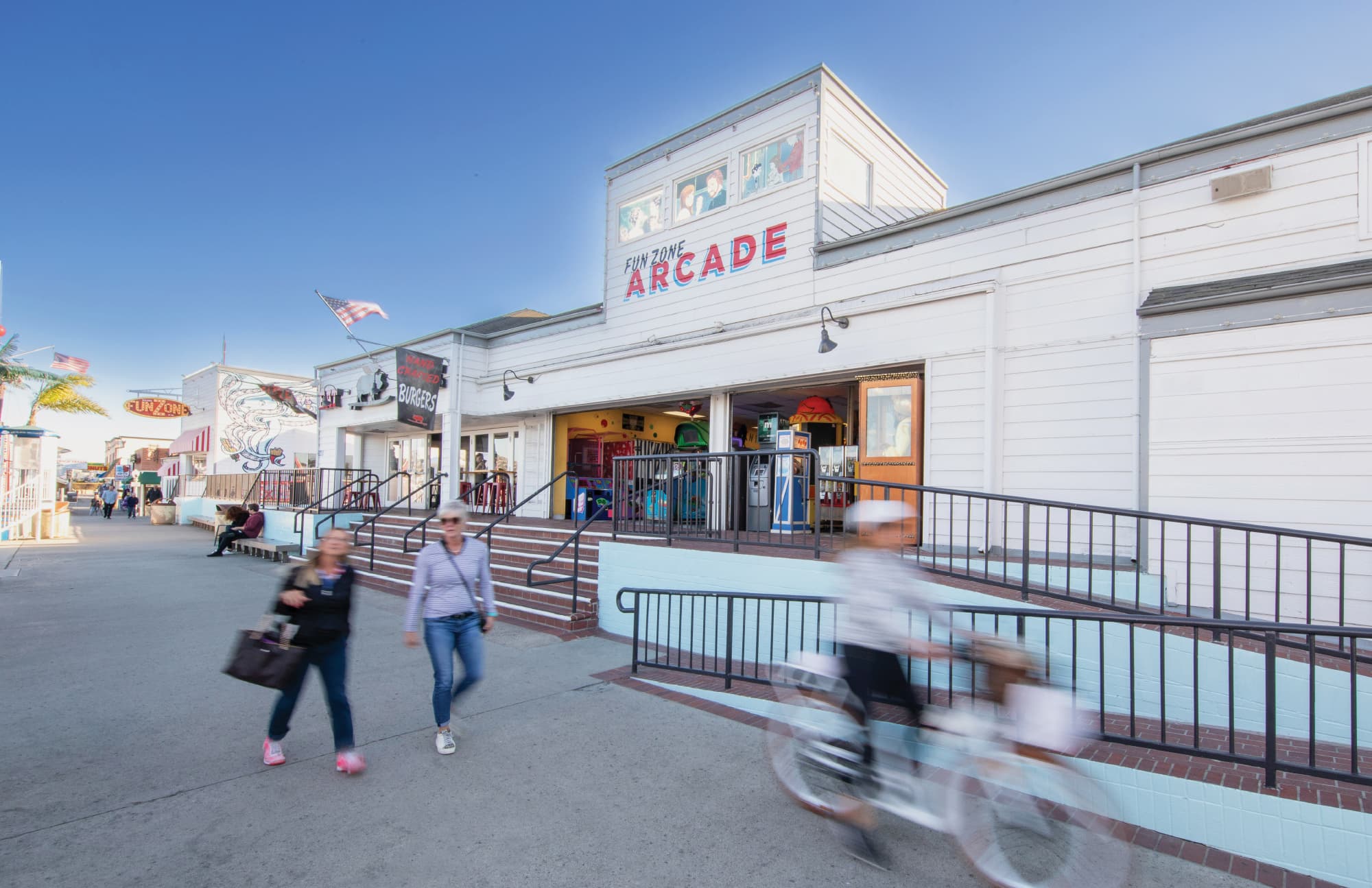 A street-level view of the Balboa Fun Zone waterfront promenade featuring the bold red and white "Fun Zone Arcade" tenant sign above an open storefront, with a burger restaurant sign, illustrated wall graphics, the Fun Zone identity sign visible down the pedestrian corridor, and visitors walking and cycling past — illustrating RSM Design's tenant signage, experiential graphics and branded environments, and placemaking services applied within the waterfront, retail, and food and beverage markets