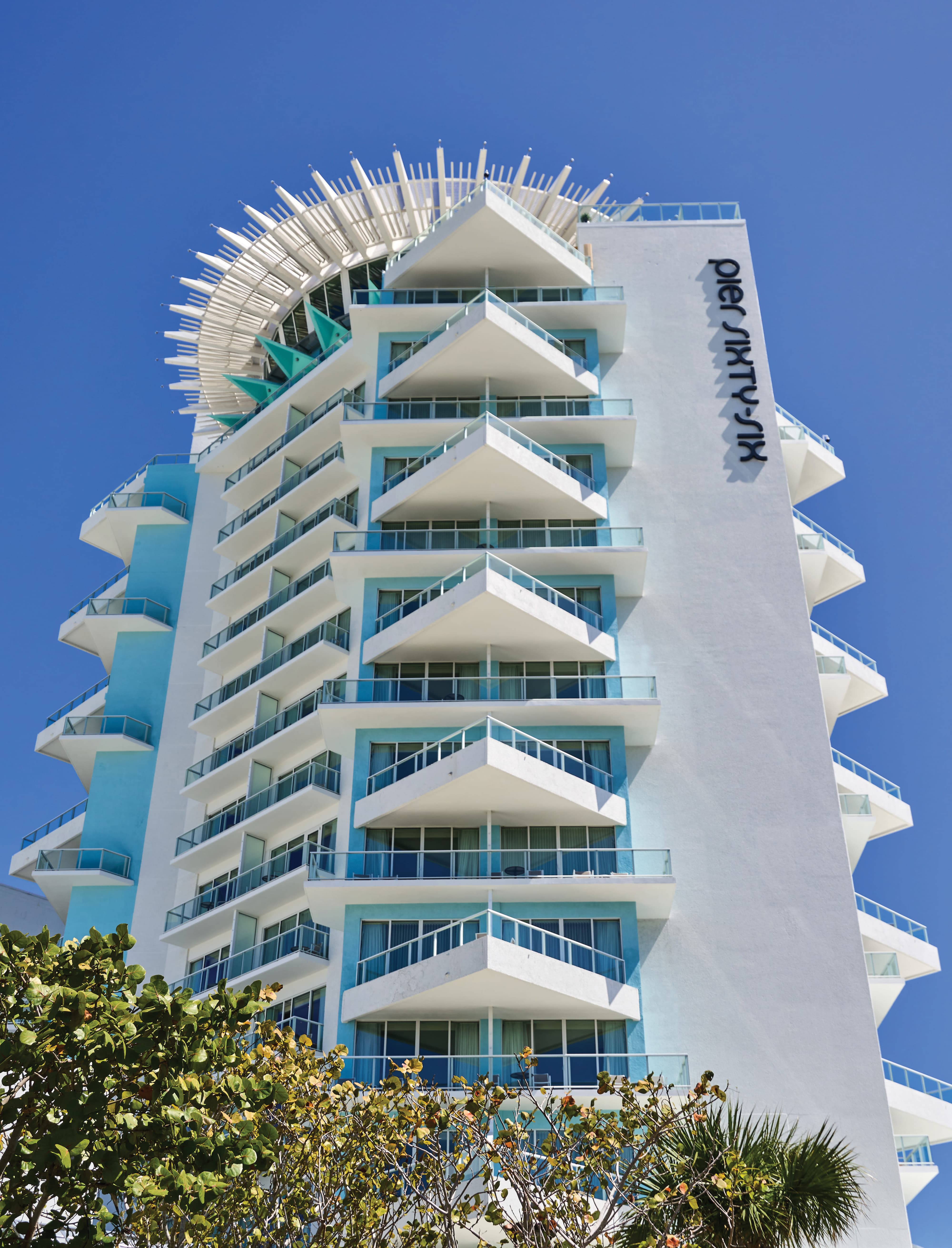 Upward view of Pier Sixty-Six luxury waterfront hotel tower in Fort Lauderdale, Florida, featuring large-scale vertical exterior building identification signage, iconic rotating rooftop structure, and staggered balconies against a blue sky — RSM Design wayfinding and signage design, master sign programs, branding and visual identity, and environmental graphic design for hospitality and waterfront destinations.