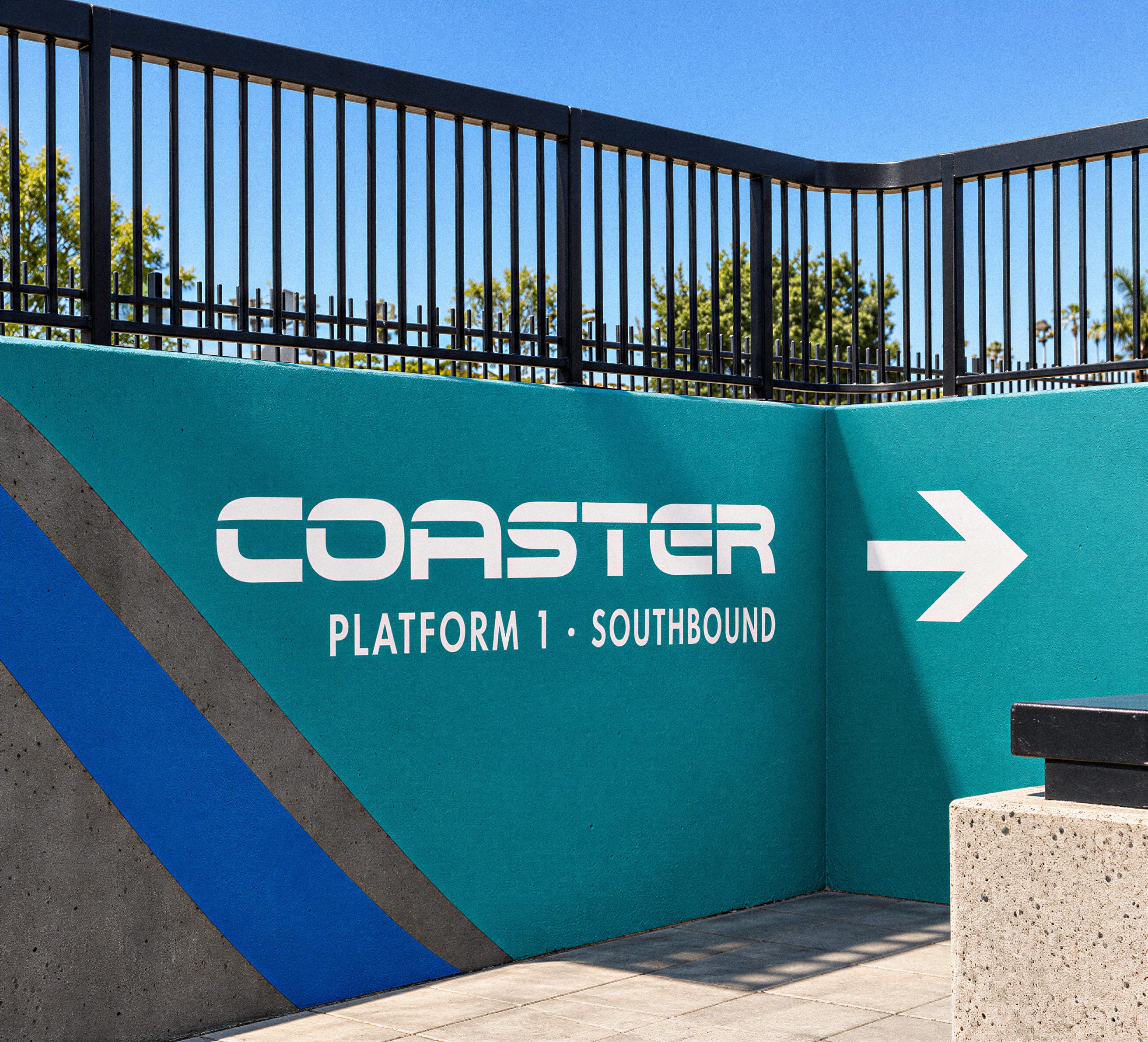 Teal wall sign reading 'Coaster, Platform 1 - Southbound' with a right-pointing arrow, mounted beneath a black metal railing at a train station.