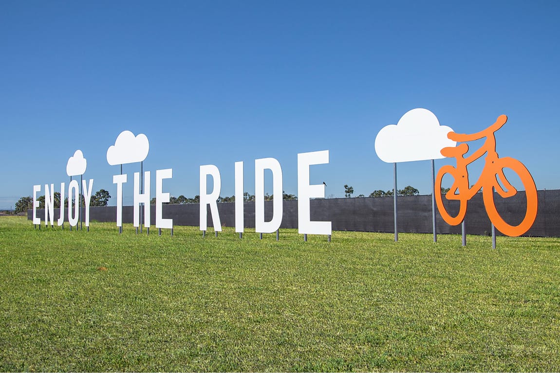Large-scale outdoor environmental signage reading "Enjoy The Ride" with white cloud cutouts and an orange bicycle sculpture installed on a green lawn, designed by RSM Design
