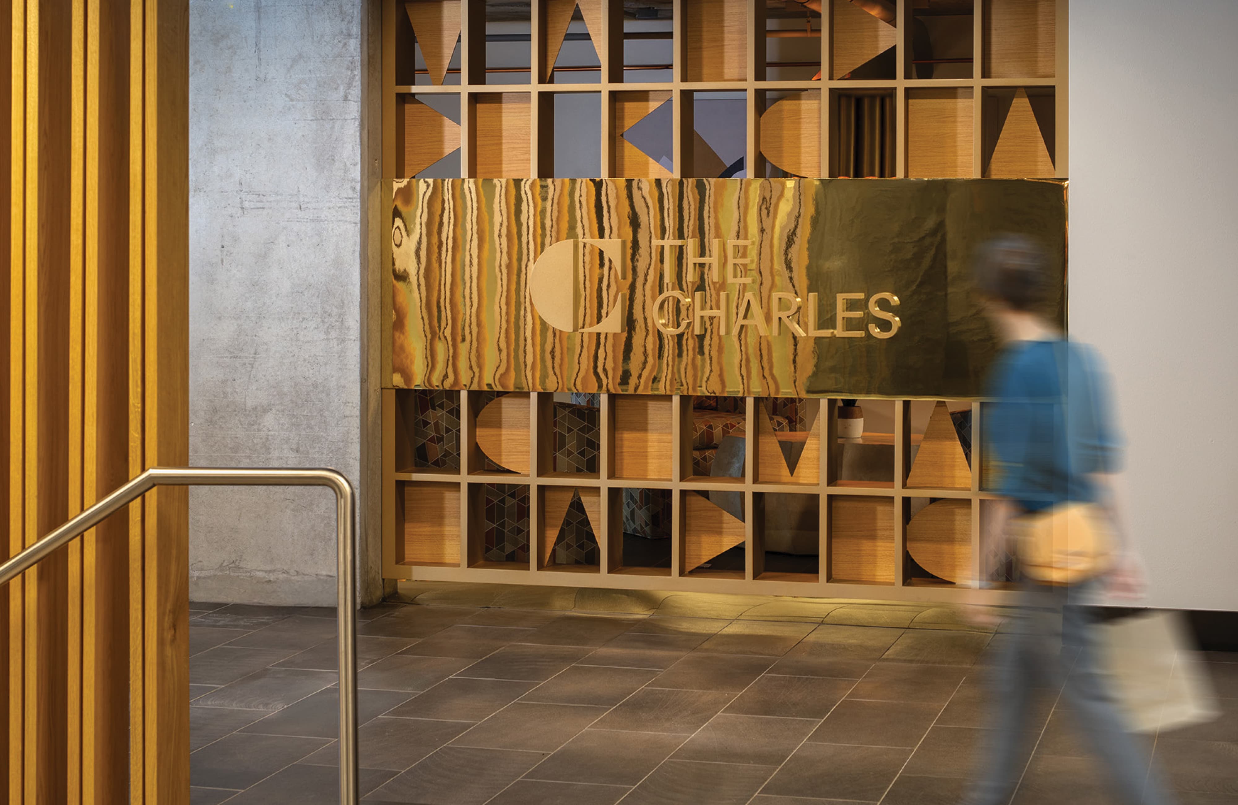 RSM Design lobby identity and experiential branded environment for The Charles at The West Quarter in Salt Lake City, featuring a floor-to-ceiling wood grid display wall filled with mixed-material geometric sculptural forms in warm tones anchored by a polished gold onyx identity panel with dimensional logo lettering, with a pedestrian descending a staircase in the foreground, demonstrating how environmental graphic design, specialty graphics, art programs, and visual identity converge with interior architecture to create a cohesive, human-centered sense of arrival for mixed-use hospitality, residential community, and experiential markets.