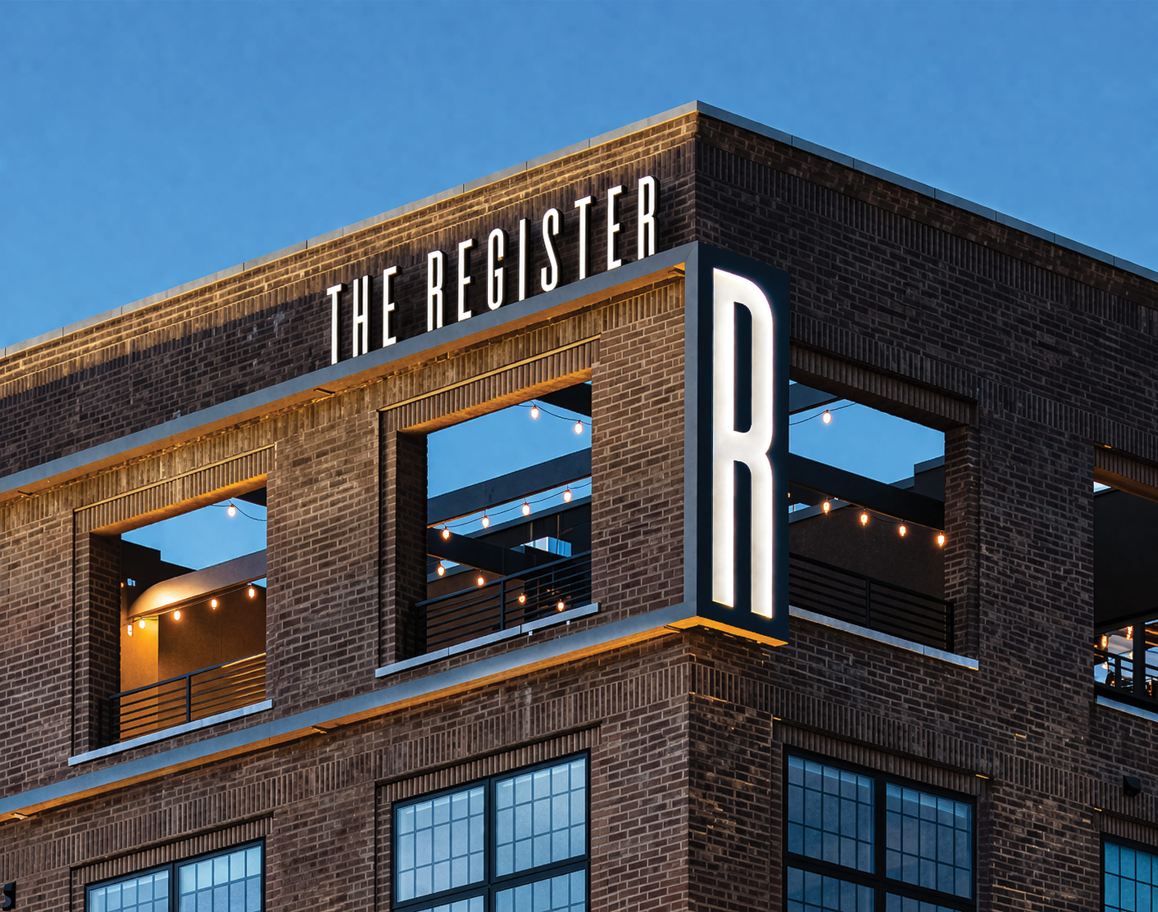 Dusk rooftop view of The Register building at Post District in Salt Lake City, featuring illuminated dimensional rooftop channel letter signage reading 'The Register' along the parapet and a large-scale backlit 'R' monogram sign on the building corner — showcasing RSM Design's architectural graphics, environmental graphic design, and branded environments for a mixed-use residential community.