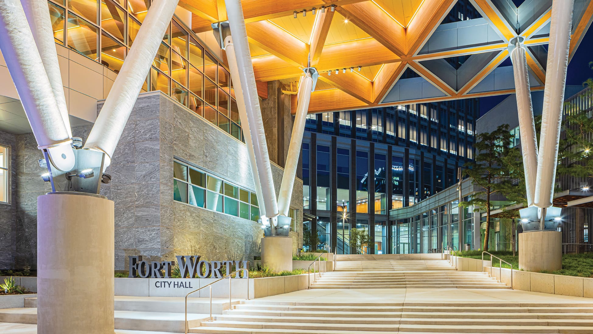 Fort Worth City Hall exterior at night featuring illuminated branching white structural columns, dramatic glowing wood lattice canopy, grand entry staircase, and dimensional City Hall monument sign on stone facade
