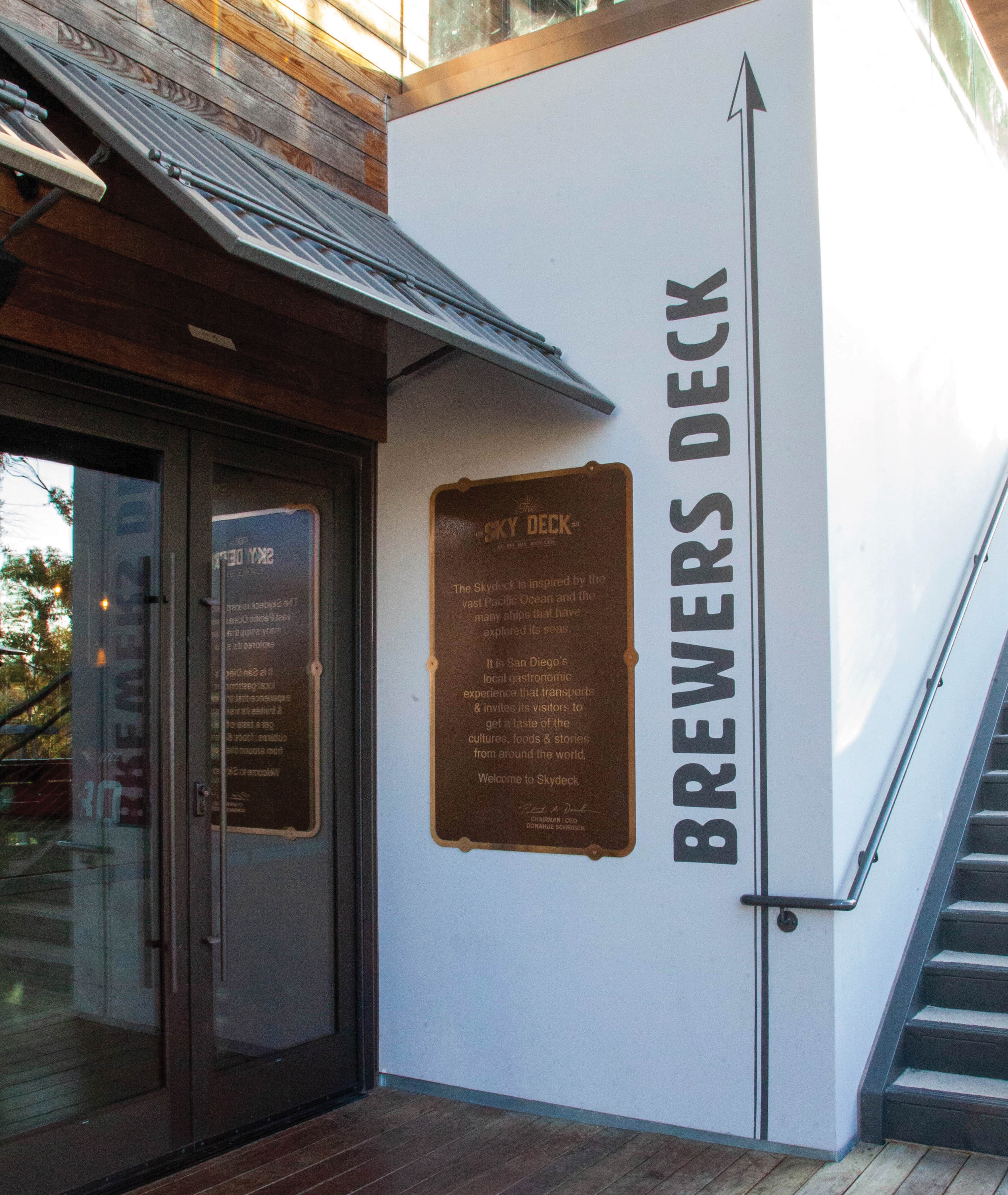 Exterior entry wall of Crafthouse Sky Deck featuring large-scale vertical "Brewers Deck" typographic wayfinding with an upward arrow, alongside a bronze interpretive plaque introducing the Sky Deck's nautical story and San Diego gastronomy concept — illustrating RSM Design's integrated approach to wayfinding and signage design, experiential graphics and branded environments, and placemaking within the food and beverage and mixed-use markets