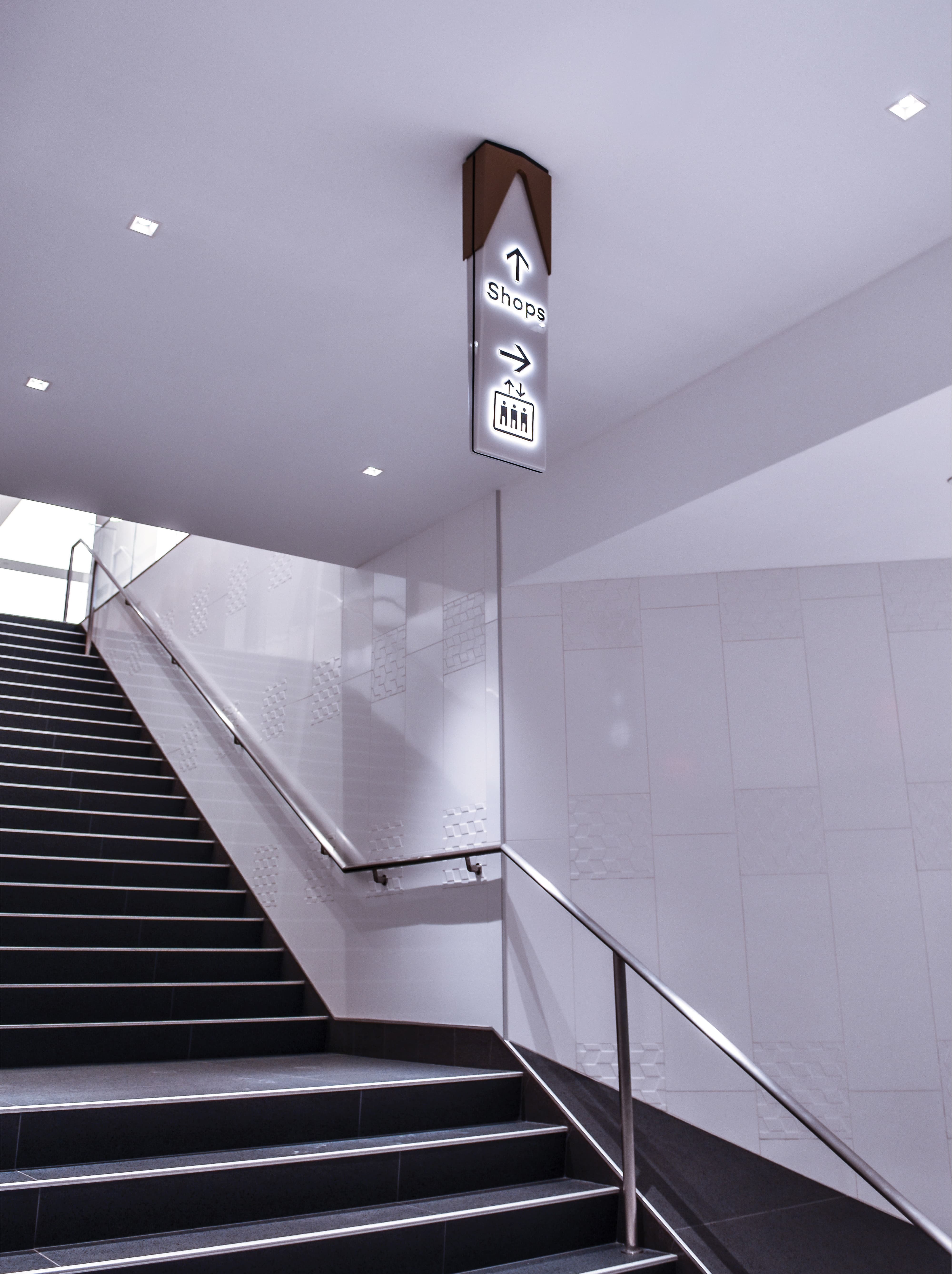 Suspended illuminated wayfinding sign directing to shops and elevators above a stairway, demonstrating environmental graphic design, signage systems, and intuitive navigation for retail, mixed-use, and commercial environments.