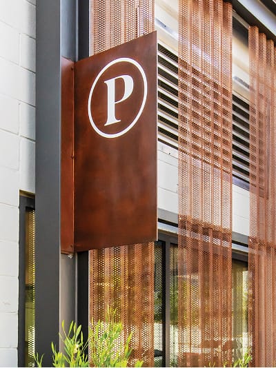 A Corten steel blade sign mounted on the exterior of a parking structure at Uncommons displays a white "P" within a circle, indicating parking. The sign is set against a perforated Corten steel screen facade, with ornamental grasses visible at the base.