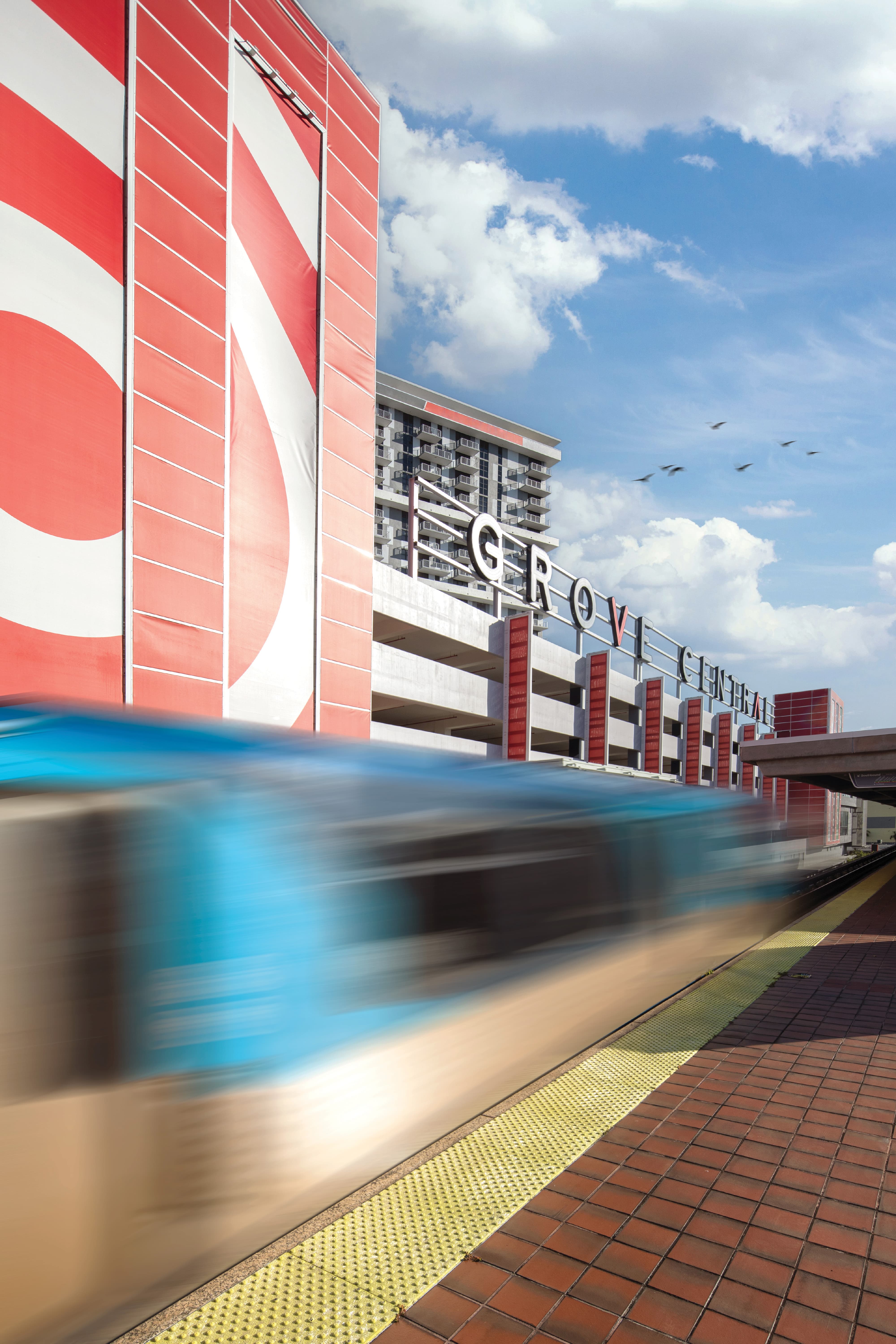 Metrorail station platform with a blurred train speeding past, set beside the red and white striped Grove Central parking structure with 'Grove Central' lettering mounted atop the building, birds flying overhead against a partly cloudy sky