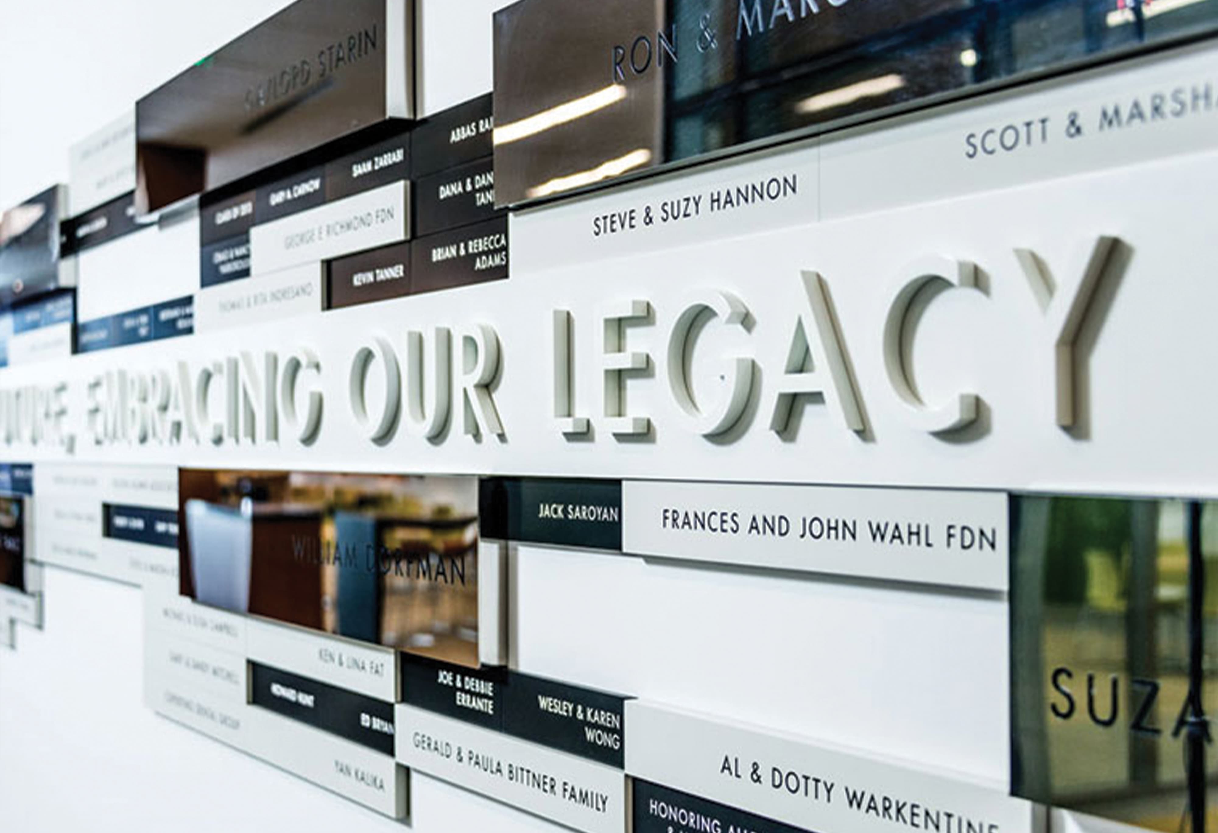 Close-up detail of a multi-tiered donor recognition wall with dimensional "Embracing Our Legacy" typographic letterforms, alternating white and dark mirror-finish donor name panels at multiple scales, at University of the Pacific Arthur A. Dugoni School of Dentistry in San Francisco — RSM Design donor recognition and specialty graphics, branded environments, and environmental graphic design for education and health and wellness destinations.