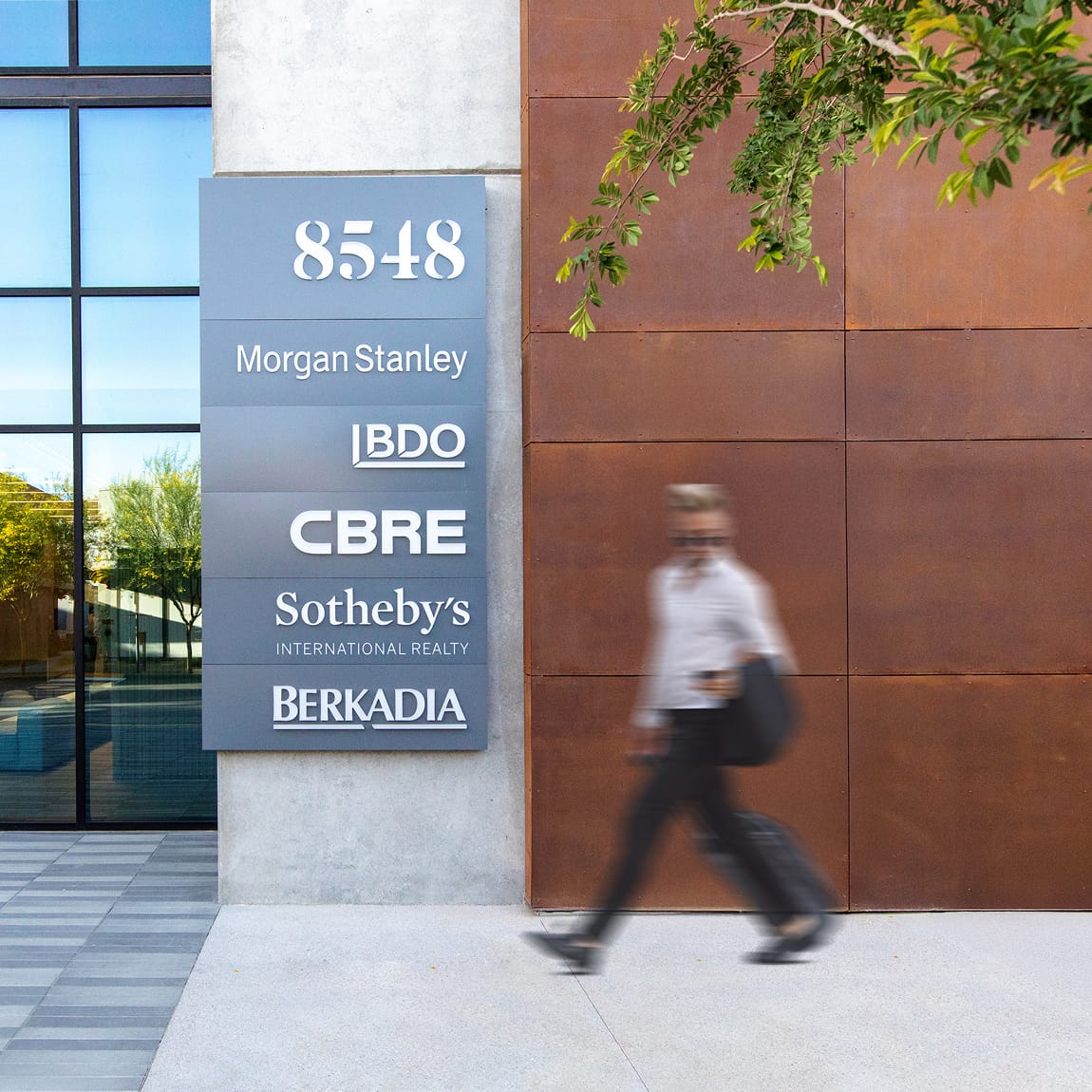 A person walks briskly past a multi-tenant directory sign mounted on the exterior of 8548 at Uncommons in Las Vegas. The gray panel sign lists tenants including Morgan Stanley, BDO, CBRE, Sotheby's International Realty, and Berkadia. The building facade features a combination of concrete, floor-to-ceiling glass, and Corten steel cladding, with a tree canopy visible above.