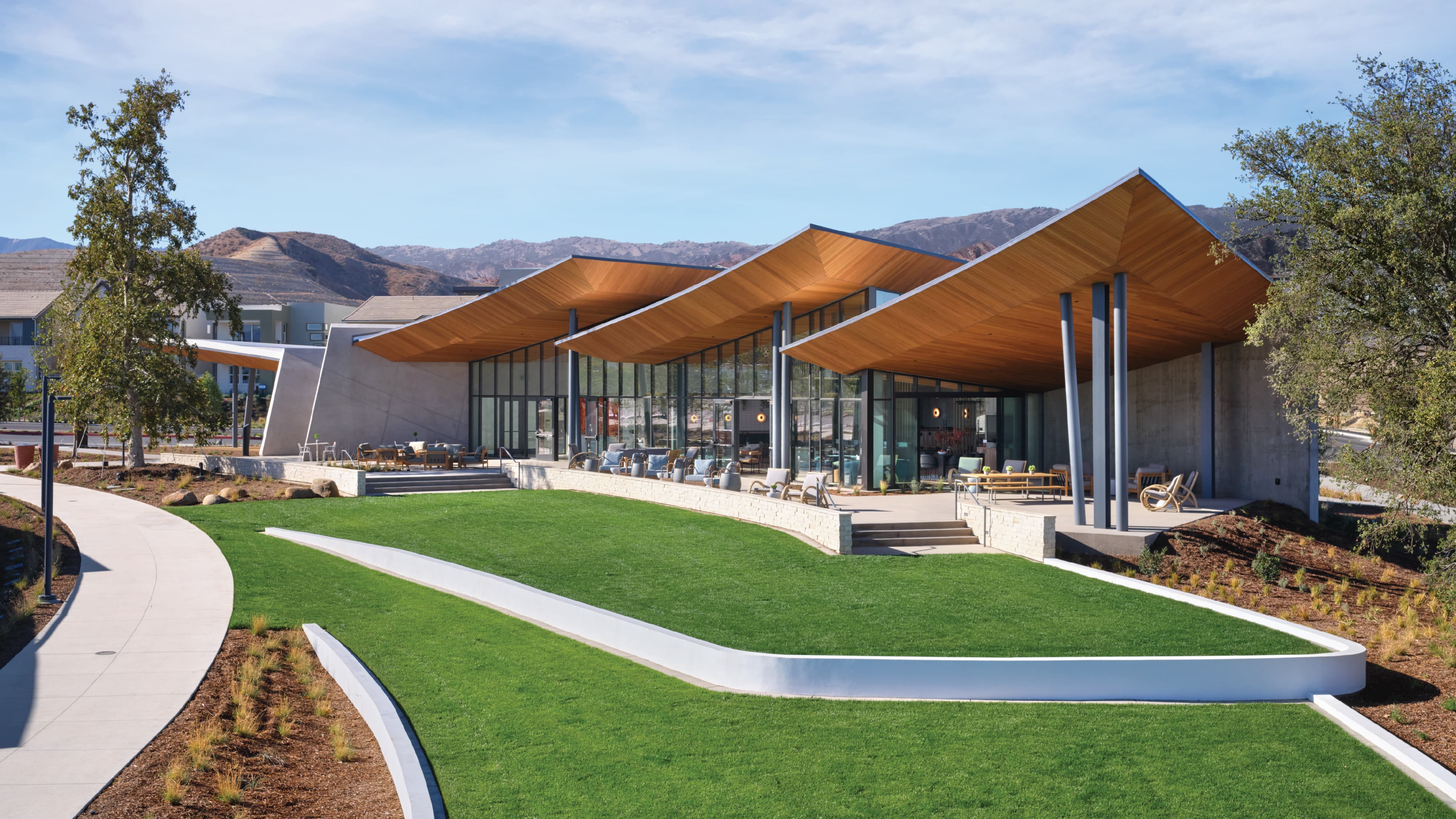 Community clubhouse with angular wood-clad roof canopies and floor-to-ceiling glass walls, overlooking a curved lawn with a walking path, set against a mountainous backdrop.