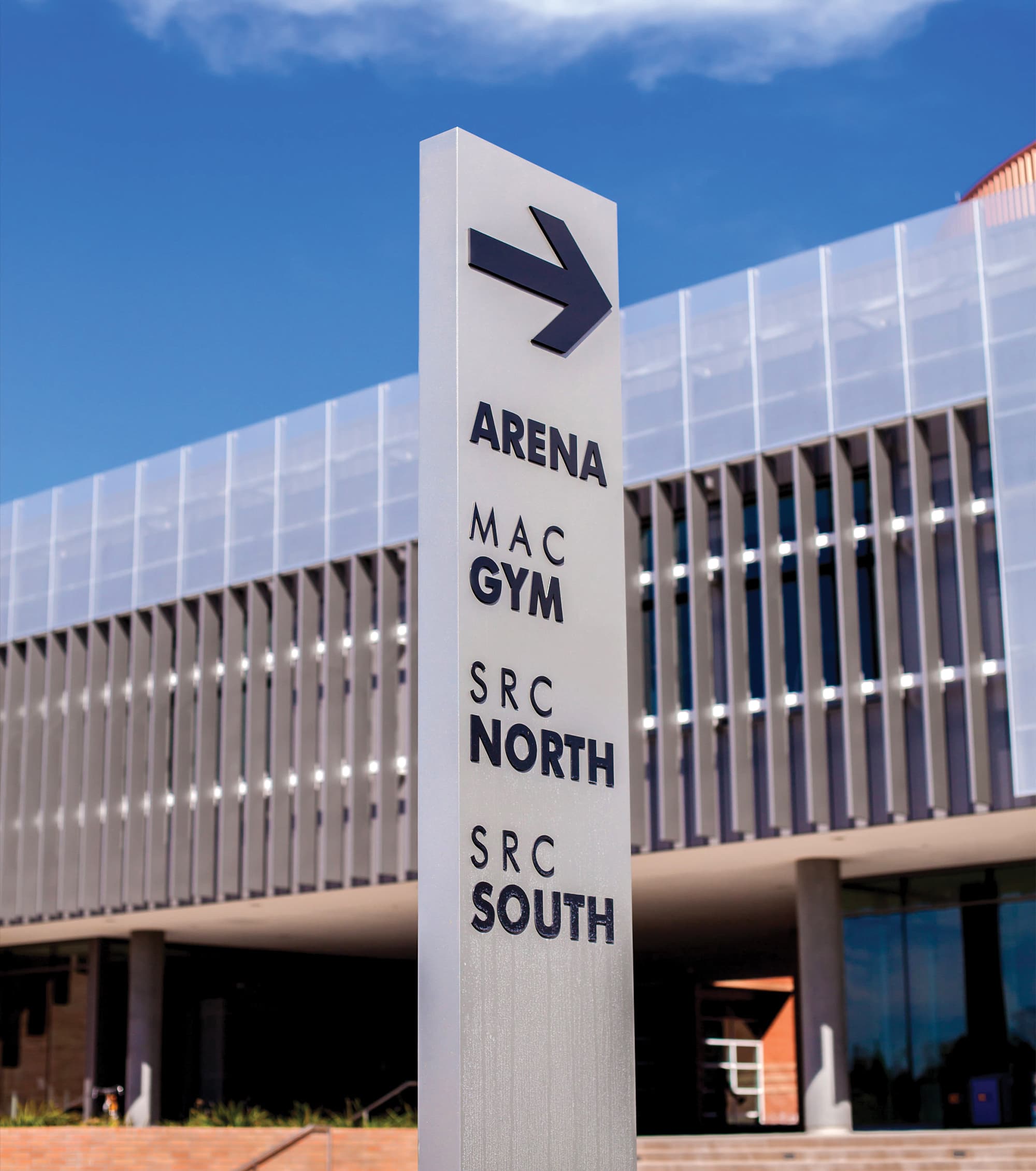 Silver directional pylon sign with a right-pointing arrow listing 'Arena, MAC Gym, SRC North, SRC South,' positioned in front of a modern building with a slatted metal facade under a blue sky.