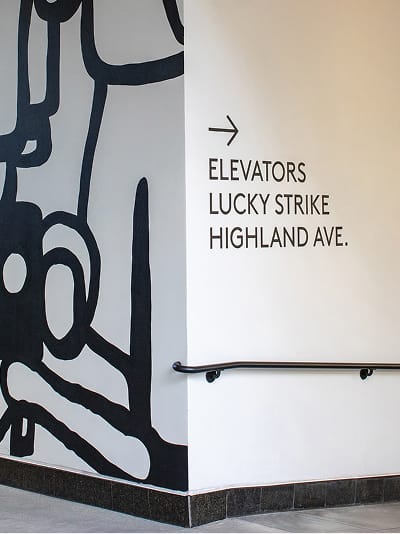 Ovation Hollywood interior directional wall signage pointing to elevators, Lucky Strike, and Highland Avenue beside bold black abstract figurative mural