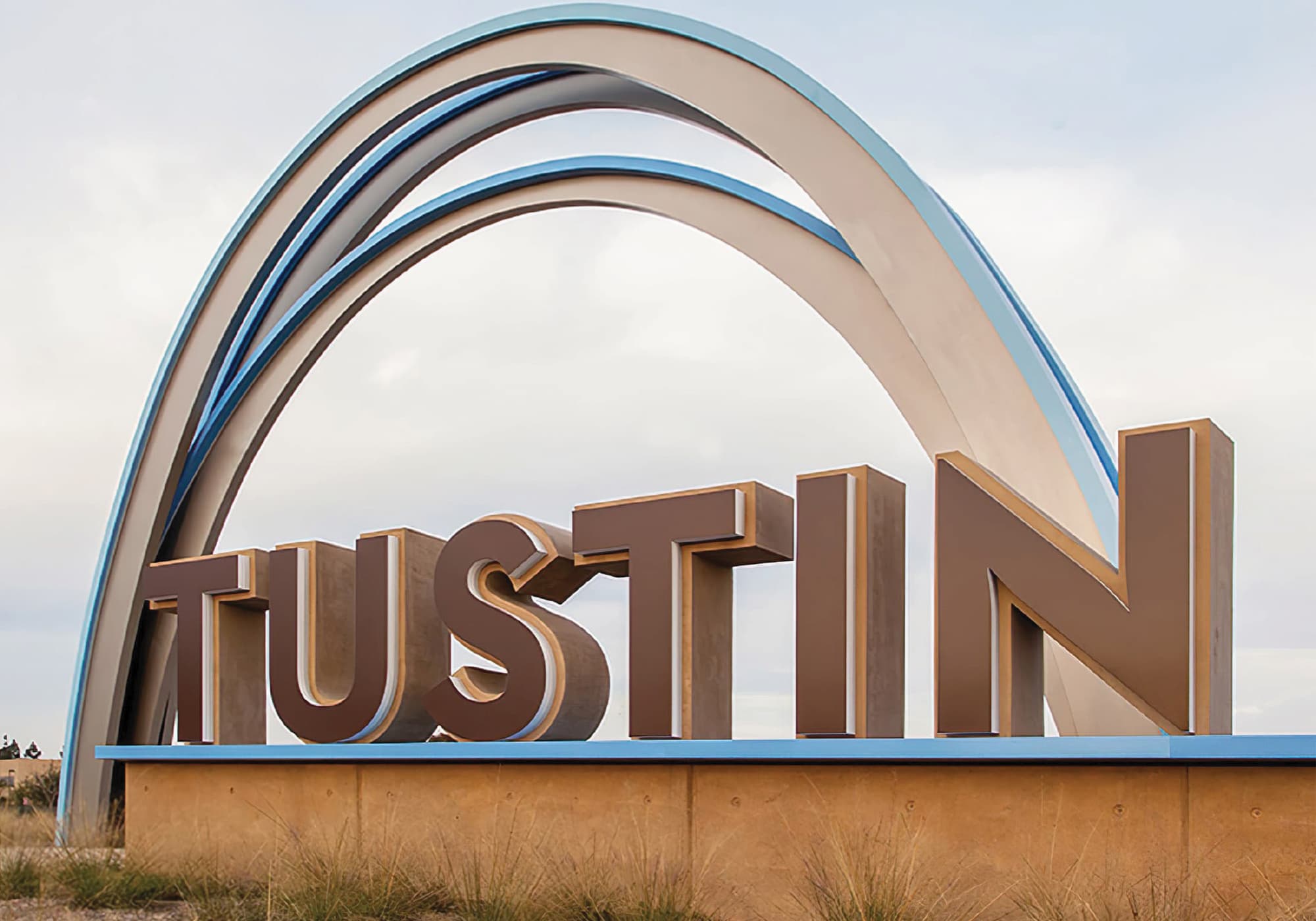 Ground-level view of large dimensional letters spelling 'Tustin' set on a stone base, framed by a curved multi-band archway rising overhead against an overcast sky, with native grasses in the foreground.