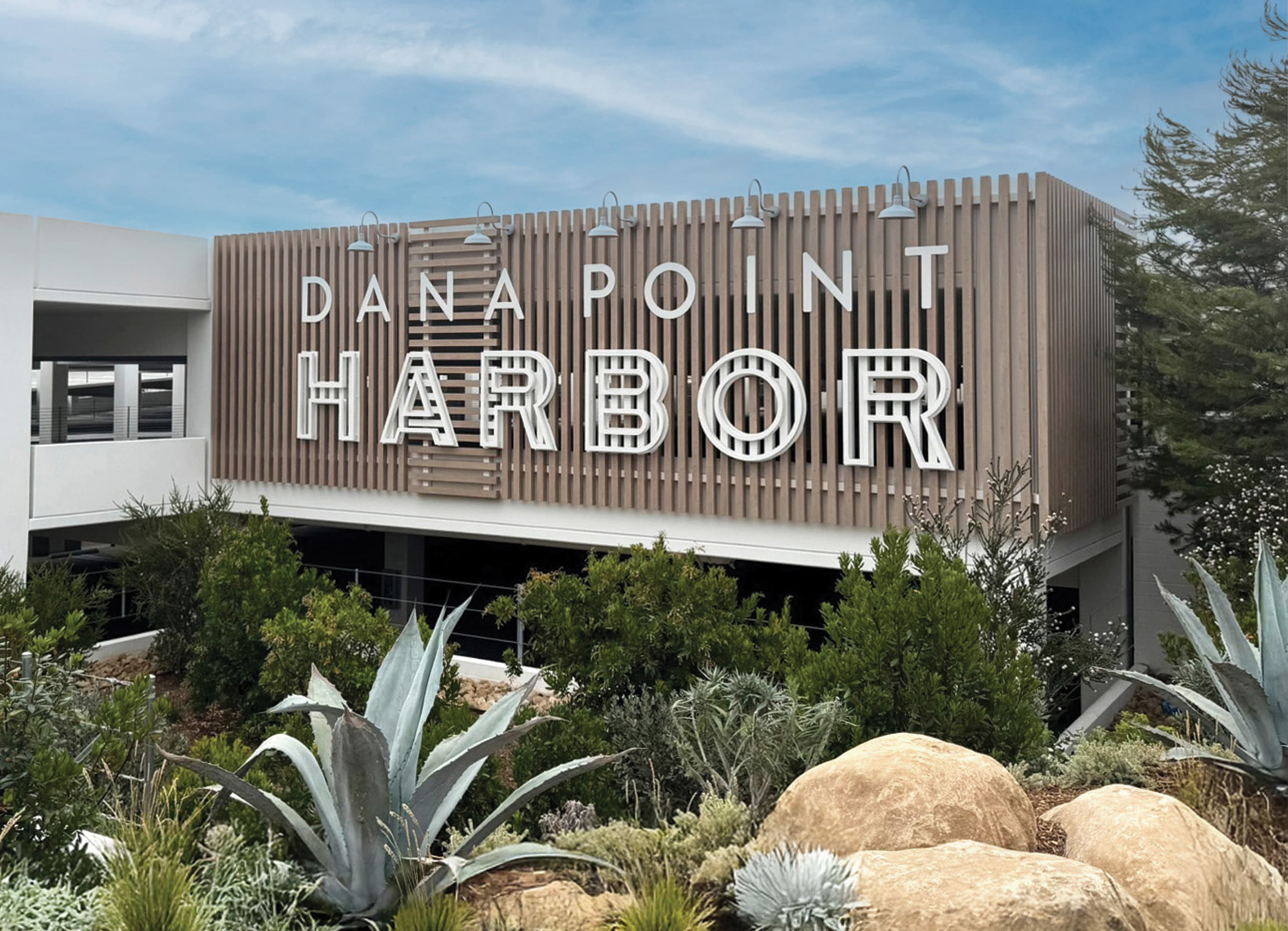 Large-scale dimensional "Dana Point Harbor" building identification sign with decorative striped letterforms mounted on a warm wood slat facade with gooseneck lighting, surrounded by native coastal succulent landscaping — RSM Design branding and visual identity, wayfinding and signage design, specialty graphics, and experiential placemaking for waterfront, civic, mixed-use, and park destinations.