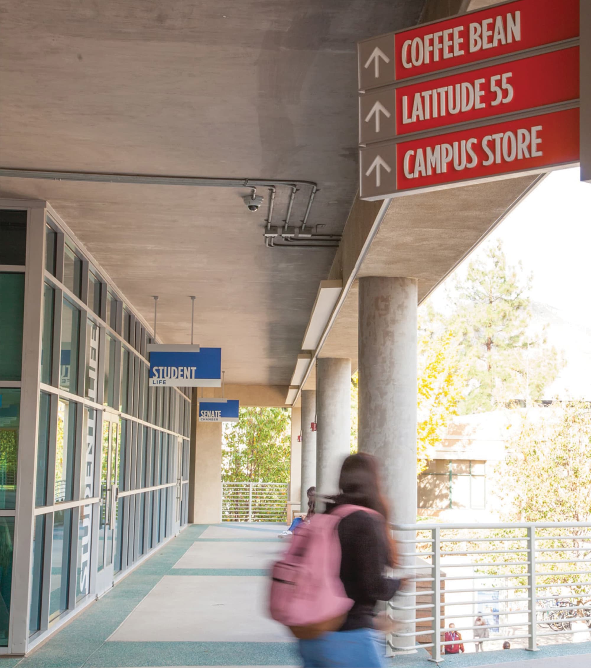 Covered campus walkway with red directional signs pointing to Coffee Bean, Latitude 55, and Campus Store, blue 'Student Life' and 'Senate Chamber' hanging signs to the left, and a student walking past in motion blur.