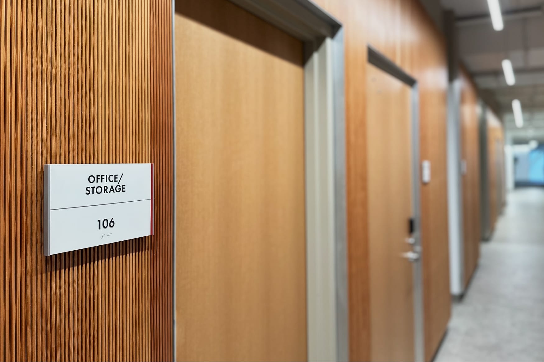 RSM Design Chapman University Rinker Campus Center Irvine — ADA-compliant room identification signage mounted on warm wood slat wall along corridor, with clean minimal typography and Braille