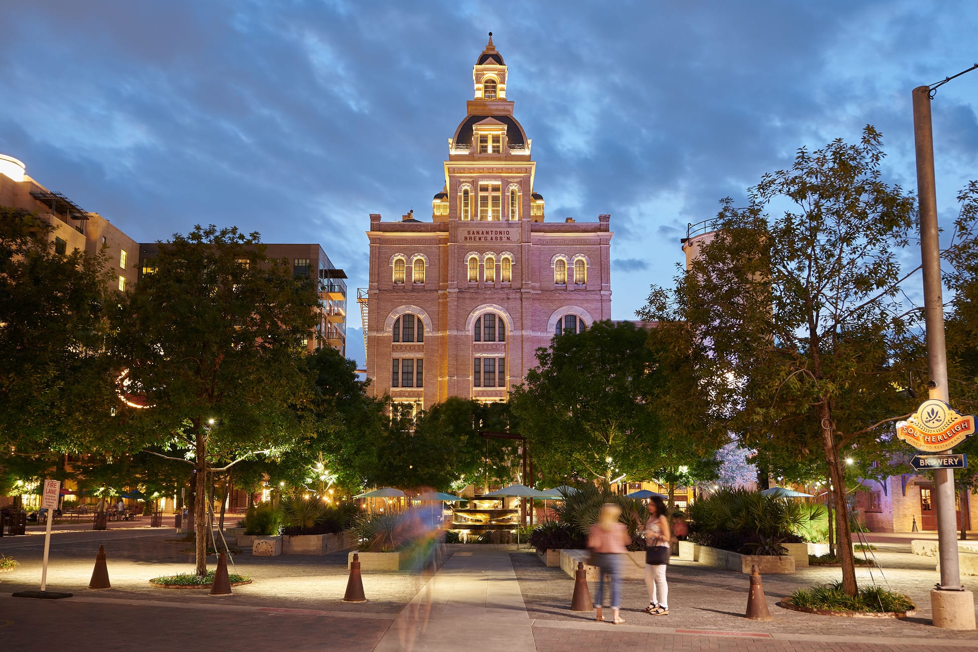 San Antonio Pearl District at night featuring the illuminated historic brewery tower, landscaped plaza, pedestrian pathways, and adaptive reuse architecture creating an active urban public space.