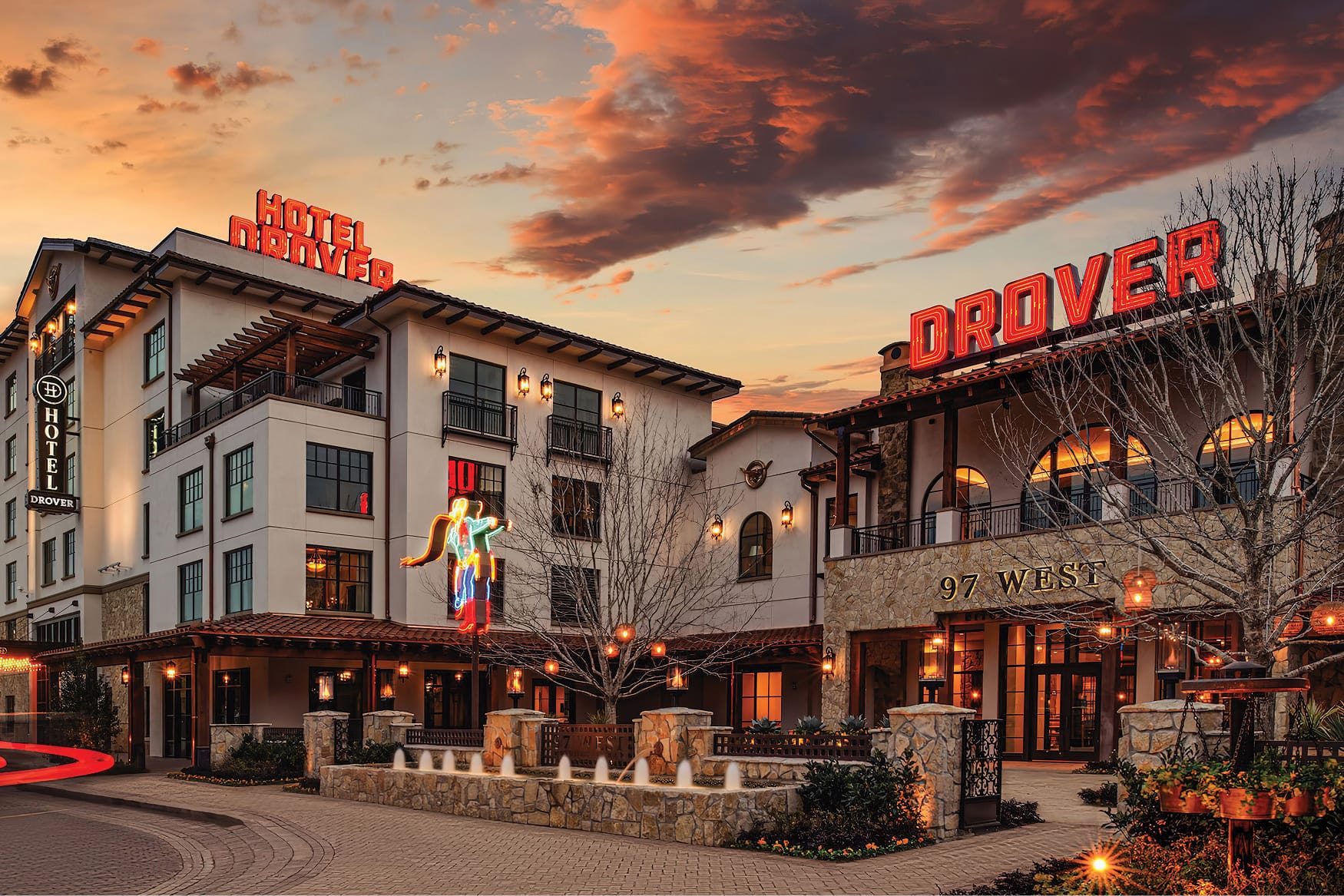 Hotel Drover exterior at sunset in Fort Worth Stockyards, Texas, featuring glowing red neon signage, a neon cowboy figure, stone and stucco facade, wrought iron lanterns, and warm ambient lighting at dusk
