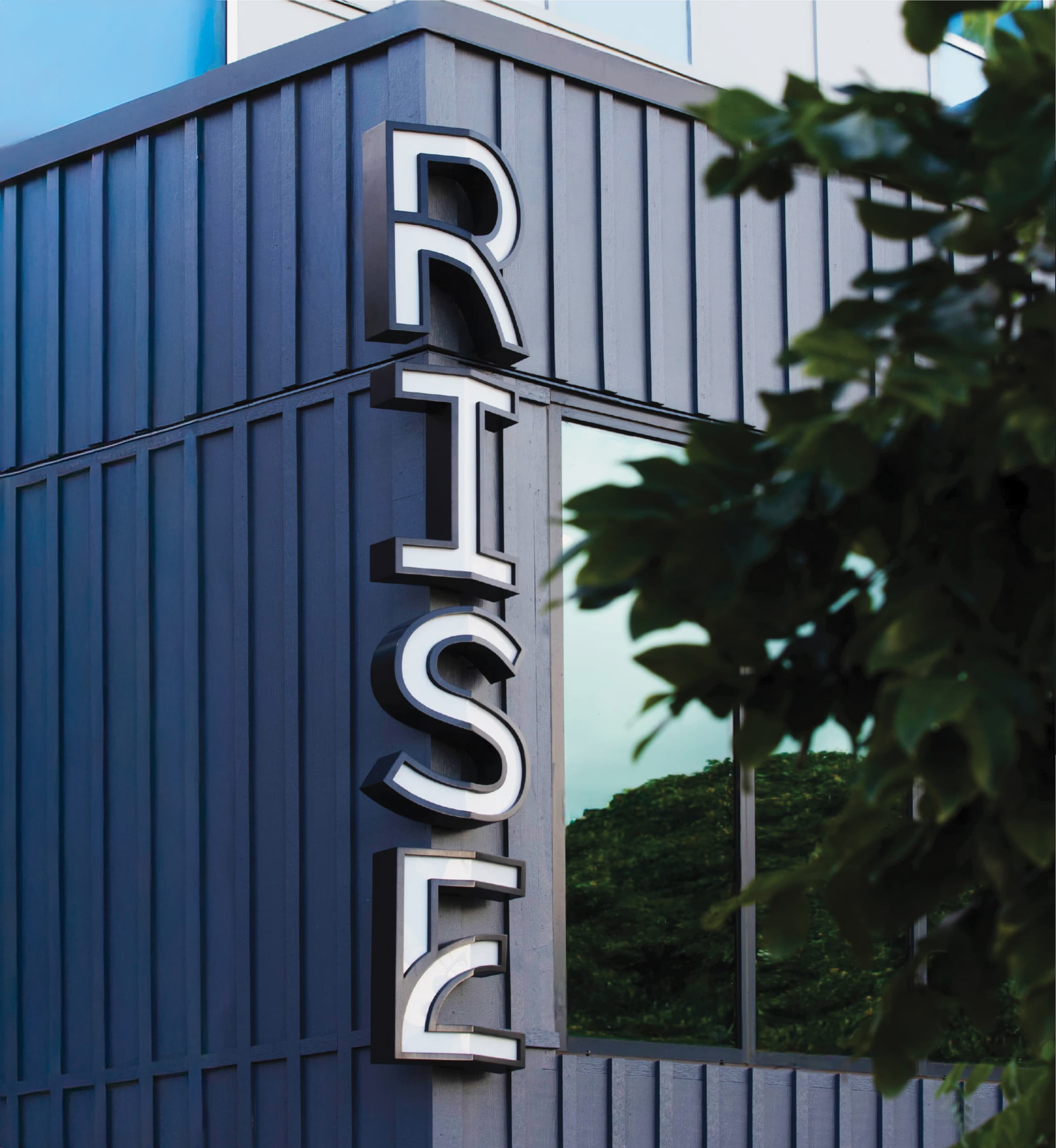Illuminated white 'RISE' letters mounted vertically on the corner of a dark gray board-and-batten building, with tree foliage in the foreground.