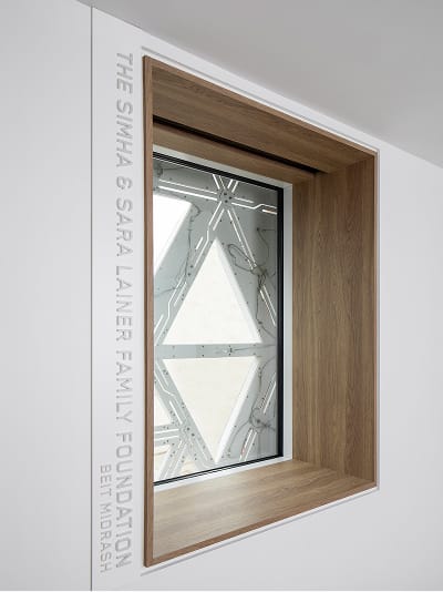 Architectural interior detail of the Simha and Sara Lainer Family Foundation Beit Midrash featuring vertical white-on-white embossed donor recognition lettering on a white wall, alongside a deep-set oak wood-framed window that frames a view of the building's exterior geometric triangular white facade panels, integrating named space signage with the architecture