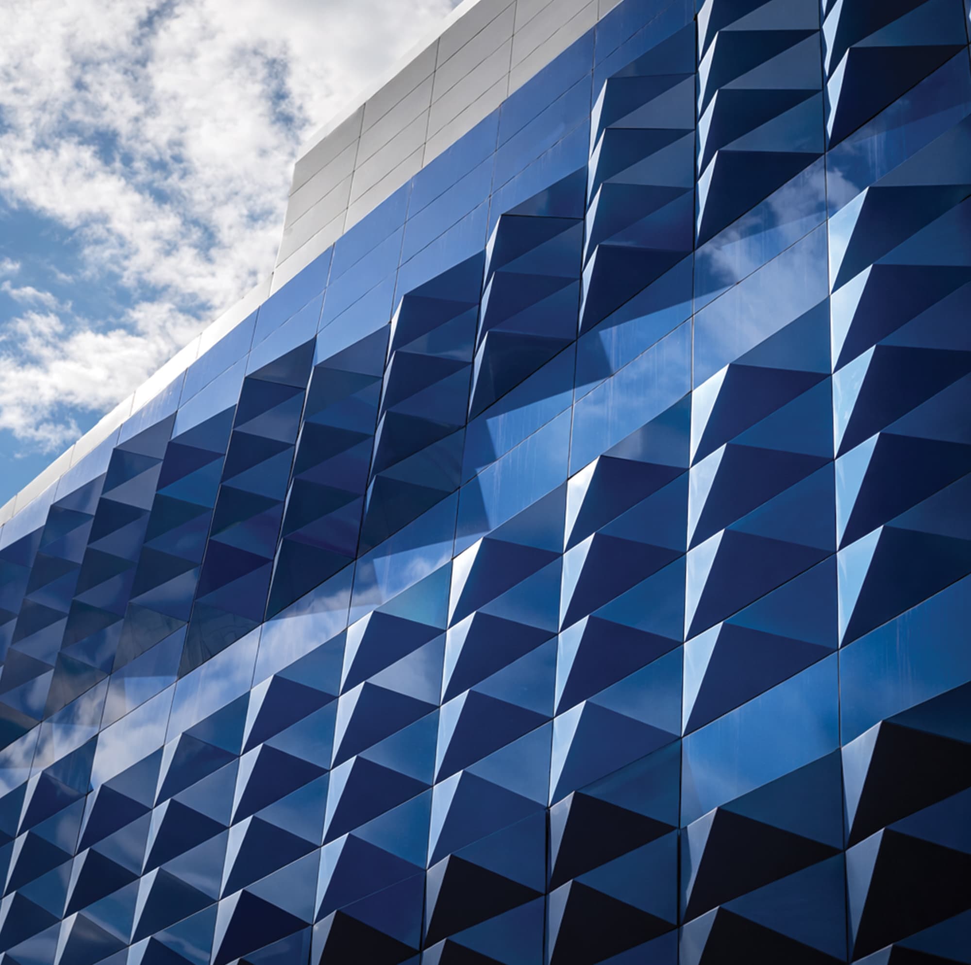 Close-up detail of a large-scale geometric triangulated blue glass exterior building facade reflecting sky and clouds at an international retail and mixed-use destination — RSM Design specialty graphics, experiential graphics, branded environments, and placemaking where environmental graphic design and architecture intersect for retail, mixed-use, and international destinations.