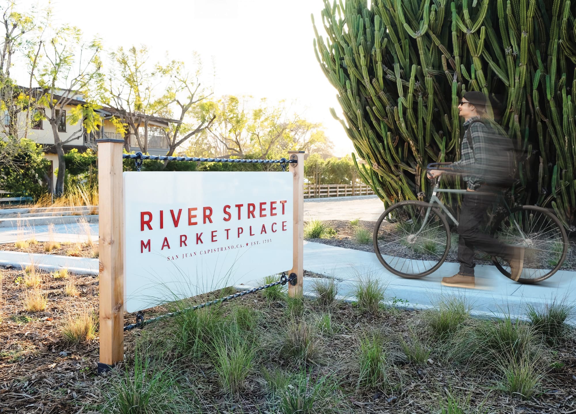 "River Street Marketplace, San Juan Capistrano, CA Est. 1794" entry identification sign suspended from wood posts with wrought iron chain hardware beside a towering cactus as a cyclist passes by, showcasing signage and placemaking design for a mixed-use waterfront retail environment by RSM Design.