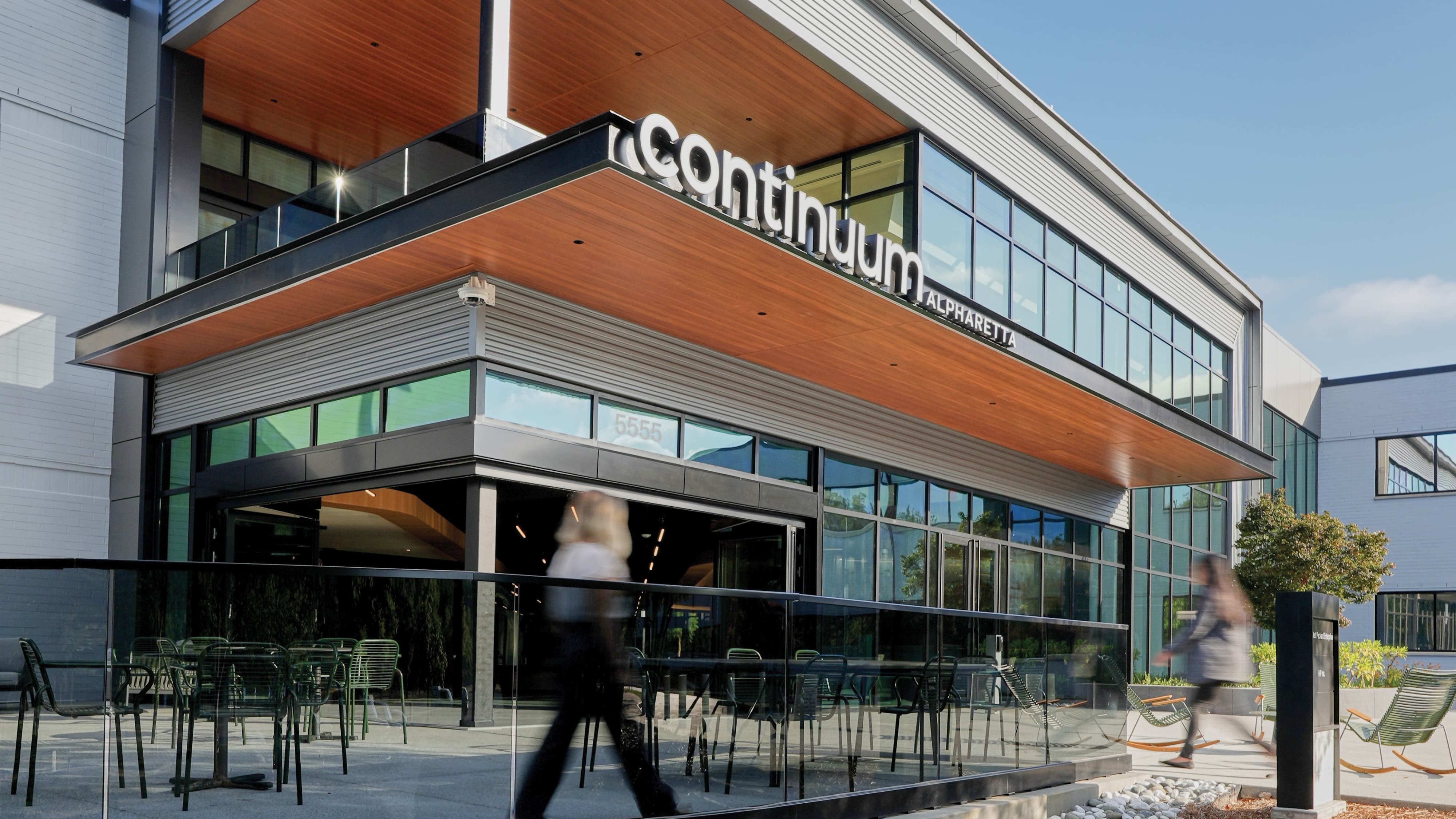 RSM Design exterior building identity signage and placemaking for Continuum Alpharetta, featuring dimensional white script and sans-serif lettering mounted on a warm wood and glass facade canopy above an activated outdoor terrace with seating, with motion-blurred pedestrians in the foreground and a branded wayfinding pylon visible at right, demonstrating how environmental graphic design, visual identity, wayfinding and signage design, and branded environments create a cohesive, human-centered sense of arrival and community for mixed-use, residential community, workplace, and health and wellness markets.