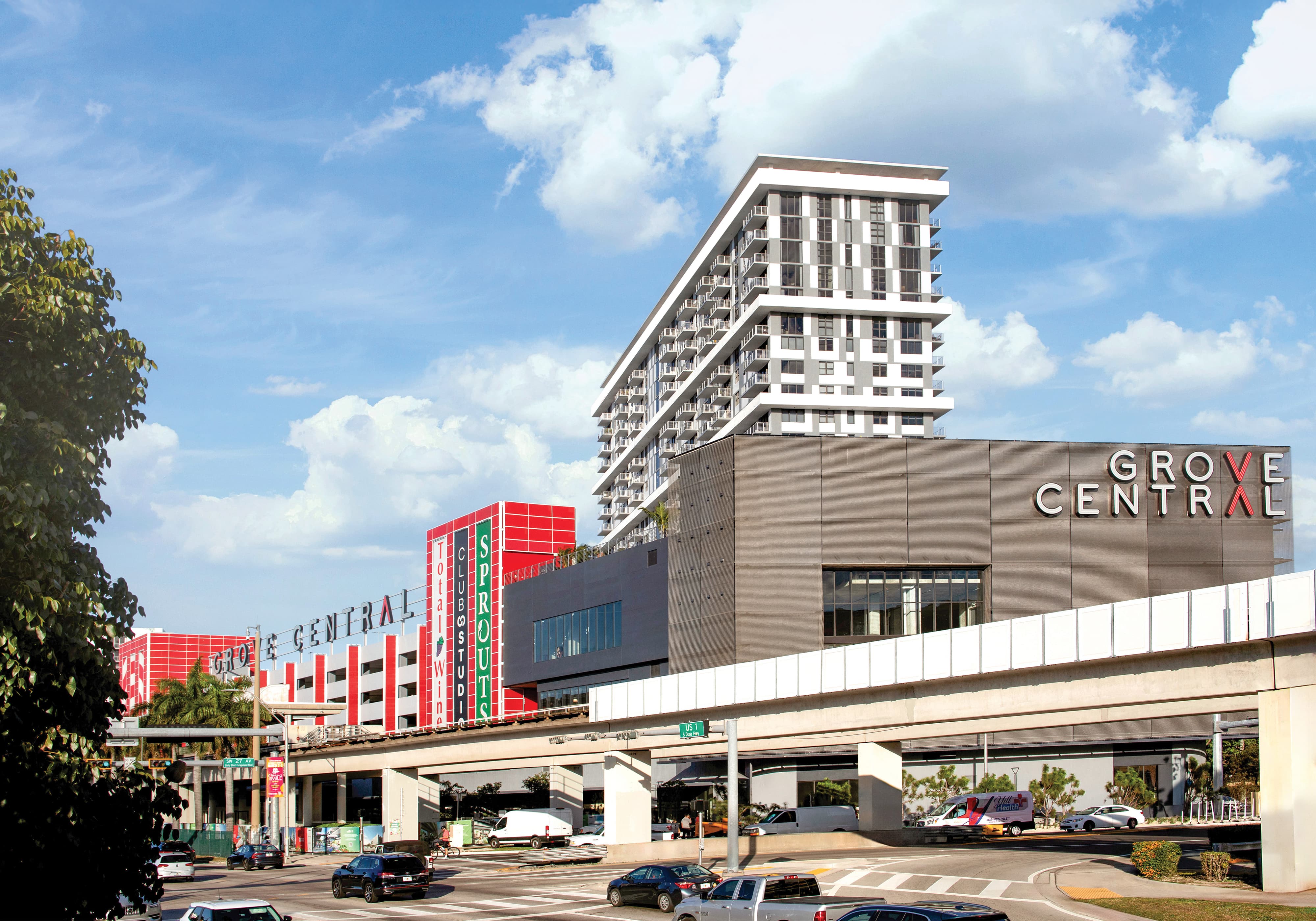 Street-level view of the Grove Central mixed-use complex, with the 'Grove Central' sign on the building facade beneath a residential tower, a red and white parking structure with retail signage for Sprouts, Club Studio, and Total Wine to the left, and an elevated Metrorail track crossing in front above a busy street intersection.