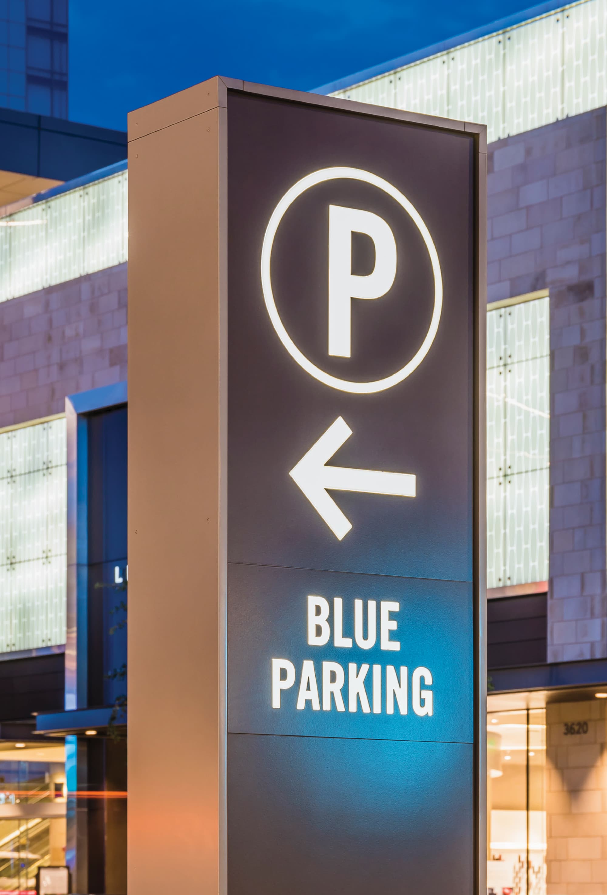 Illuminated exterior wayfinding pylon sign directing visitors to Blue Parking at a mixed-use retail destination, featuring a backlit parking symbol and directional arrow — part of an integrated wayfinding signage system designed by RSM Design for a parking garage and retail environment.