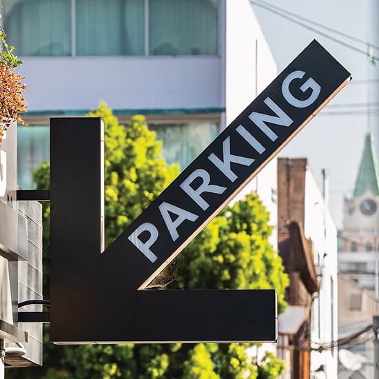 Bold black diagonal arrow parking directional sign pointing down at Jack London Square with historic clock tower and urban streetscape in background, Oakland, California