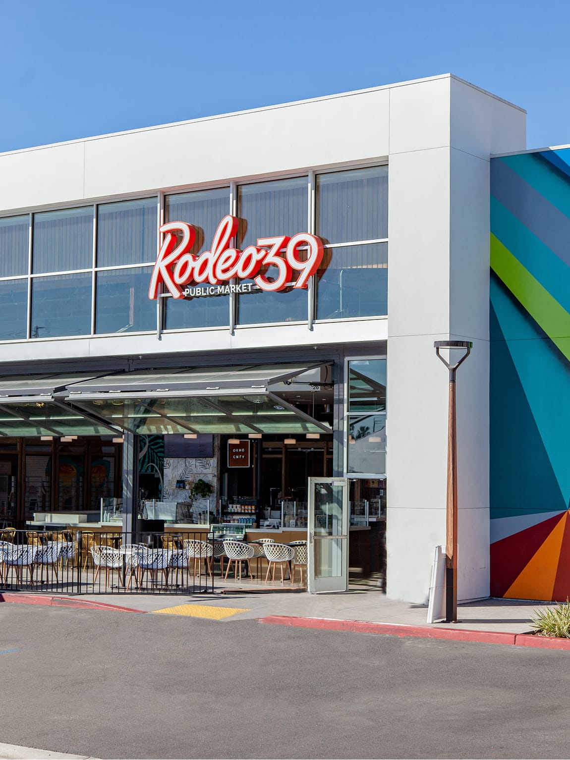 Daytime angled exterior view of Rodeo 39 Public Market in Stanton, CA, showcasing the bold red neon sign above floor-to-ceiling glazing, open patio seating with white chairs, and the edge of the vivid geometric mural in teal, green, orange, and red visible on the adjacent facade.