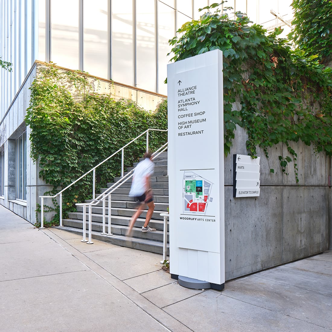 Pedestrian directional sign with campus map listing the Alliance Theatre, Atlanta Symphony Hall, and High Museum of Art, beside a MARTA transit blade sign and outdoor stairway with ivy-covered walls at the Woodruff Arts Center – campus wayfinding and environmental graphic design by RSM Design.