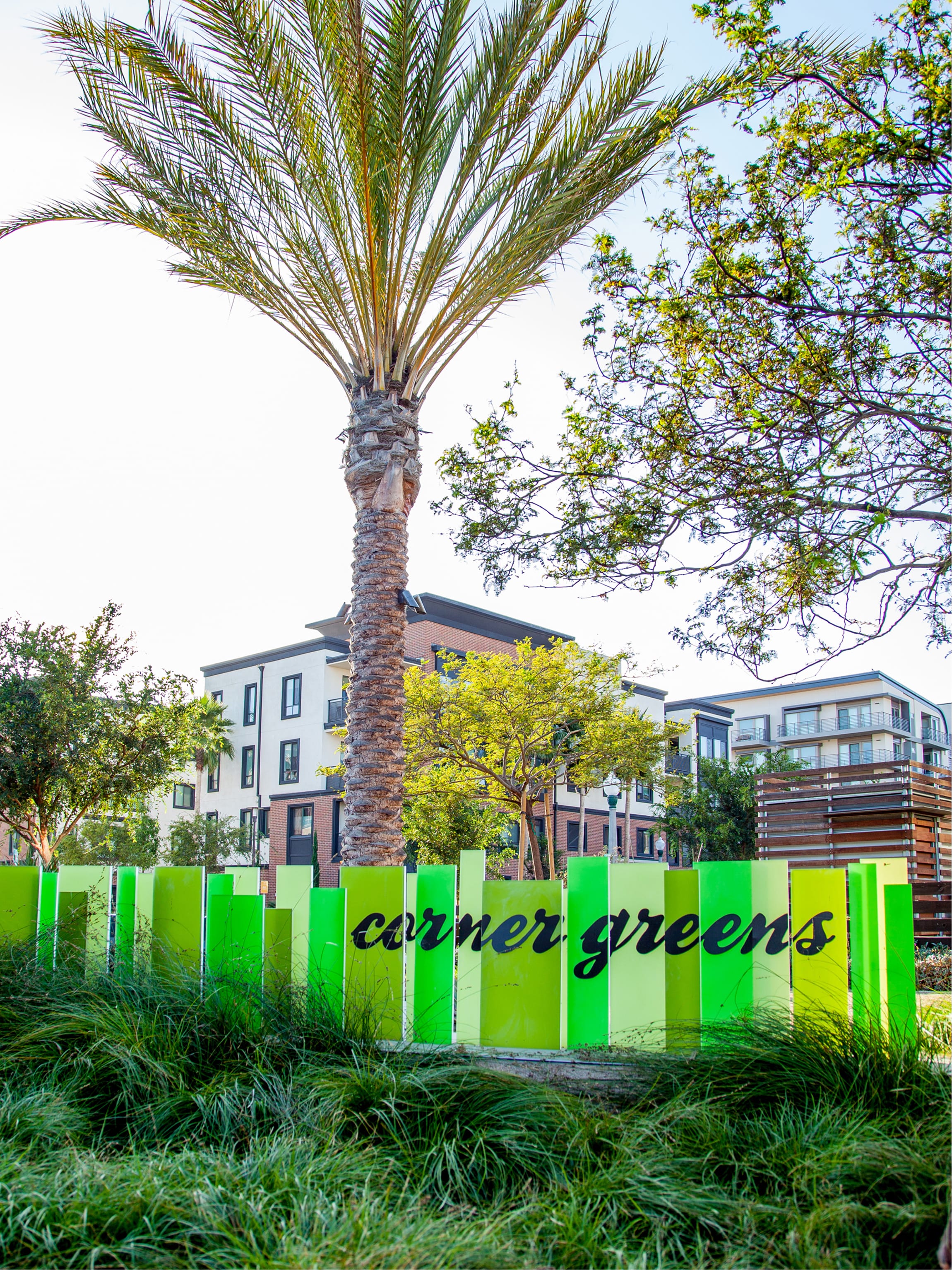 Vertical green panel sign spelling "corner greens" in script lettering, designed by RSM Design, set among tall grasses with a palm tree and residential buildings in the background at Playa Vista.