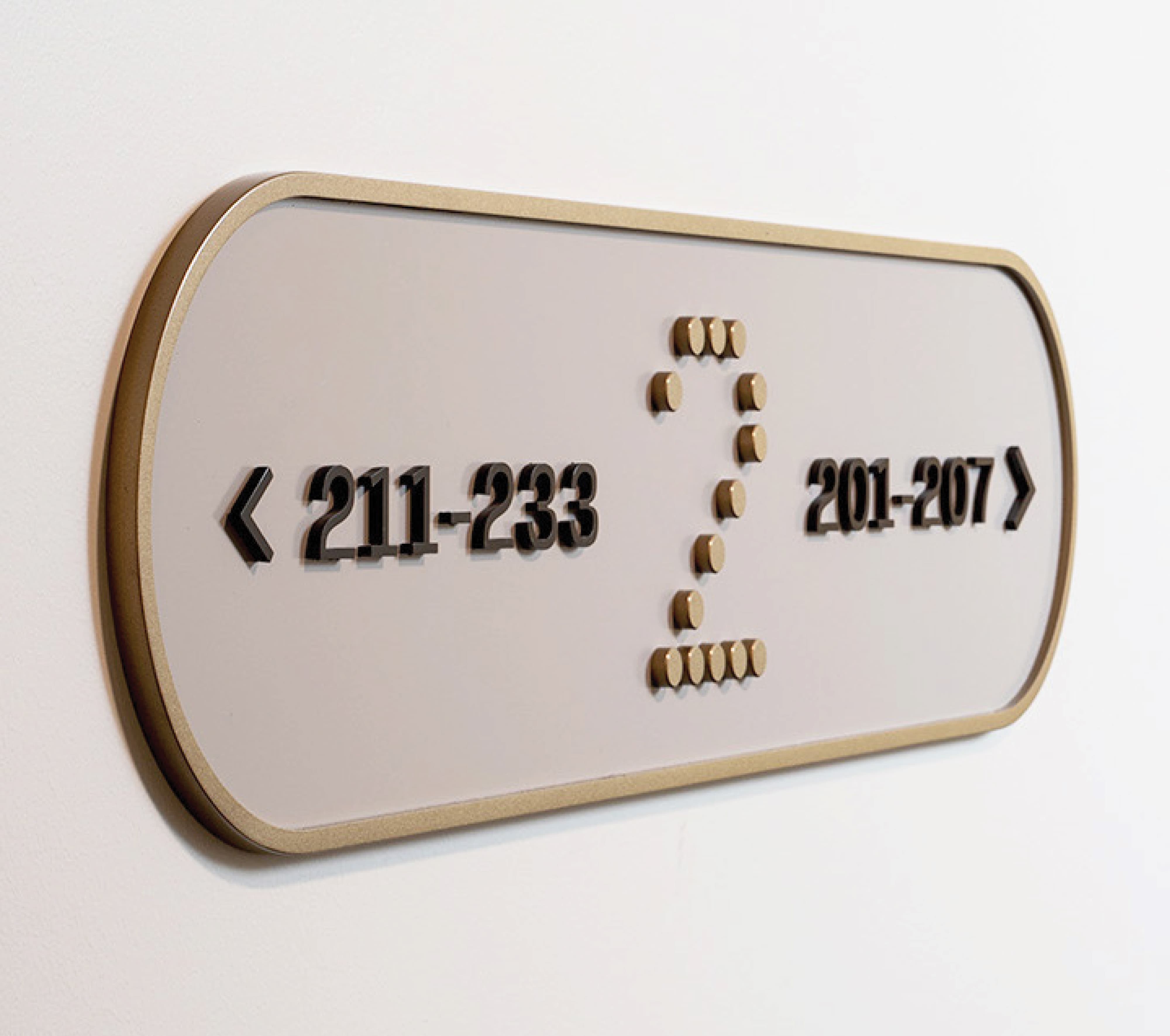 RSM Design interior corridor wayfinding and ADA-compliant signage for The Charles at The West Quarter in Salt Lake City, featuring a close-up of a cream pill-shaped directional plaque with dimensional black unit range numerals, gold chevron arrows, and tactile Braille indicators set within a gold glitter edge frame, demonstrating how wayfinding and signage design, specialty graphics, environmental graphic design, and master sign program execution integrate accessibility compliance seamlessly into a cohesive, refined branded environment for mixed-use hospitality and residential community markets.