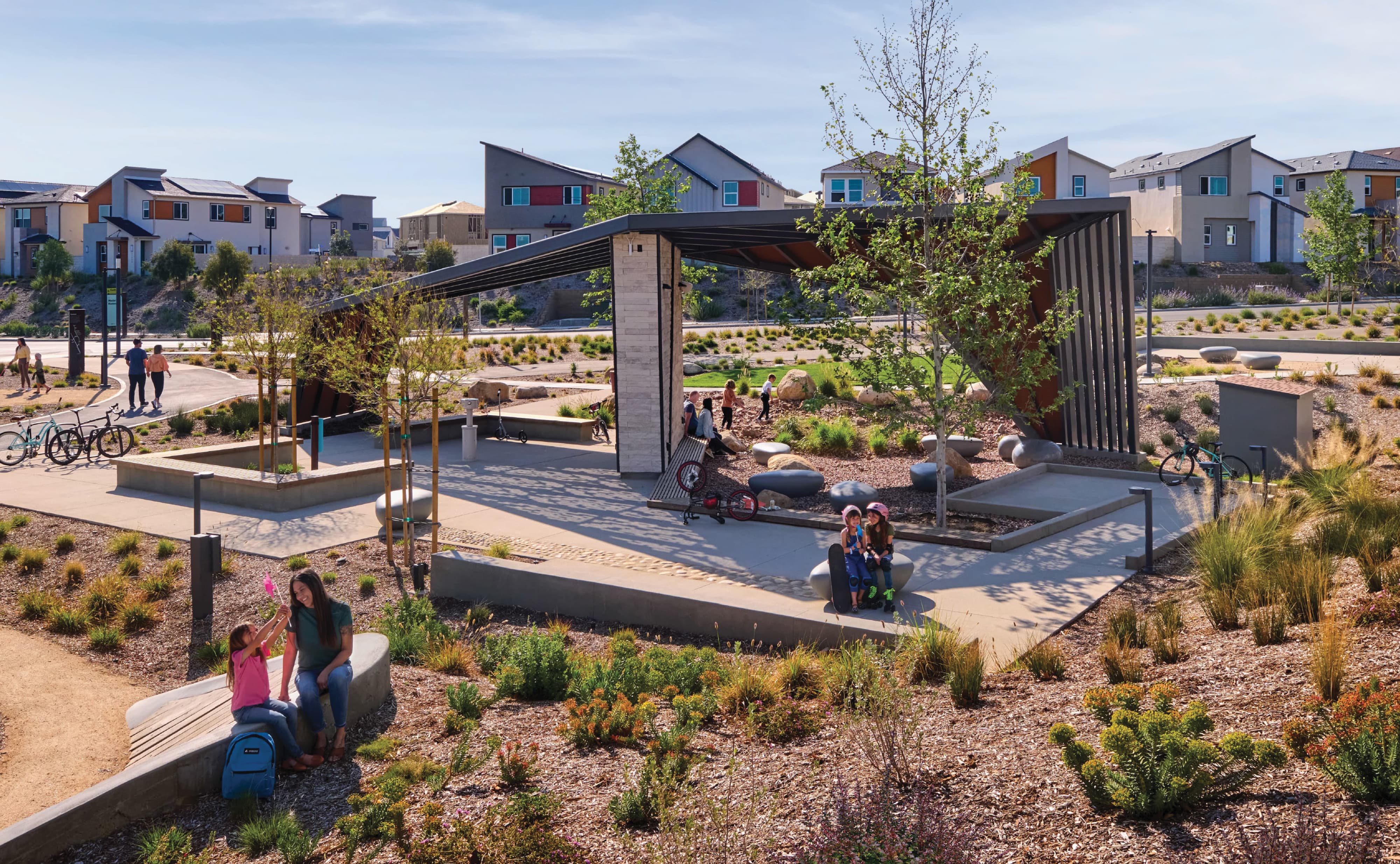 Community trailhead pavilion with an angular metal roof and stone column, surrounded by boulder seating, bike racks, and native landscaping, with children on bikes and skateboards, a parent and child on a bench, and residential homes in the background.
