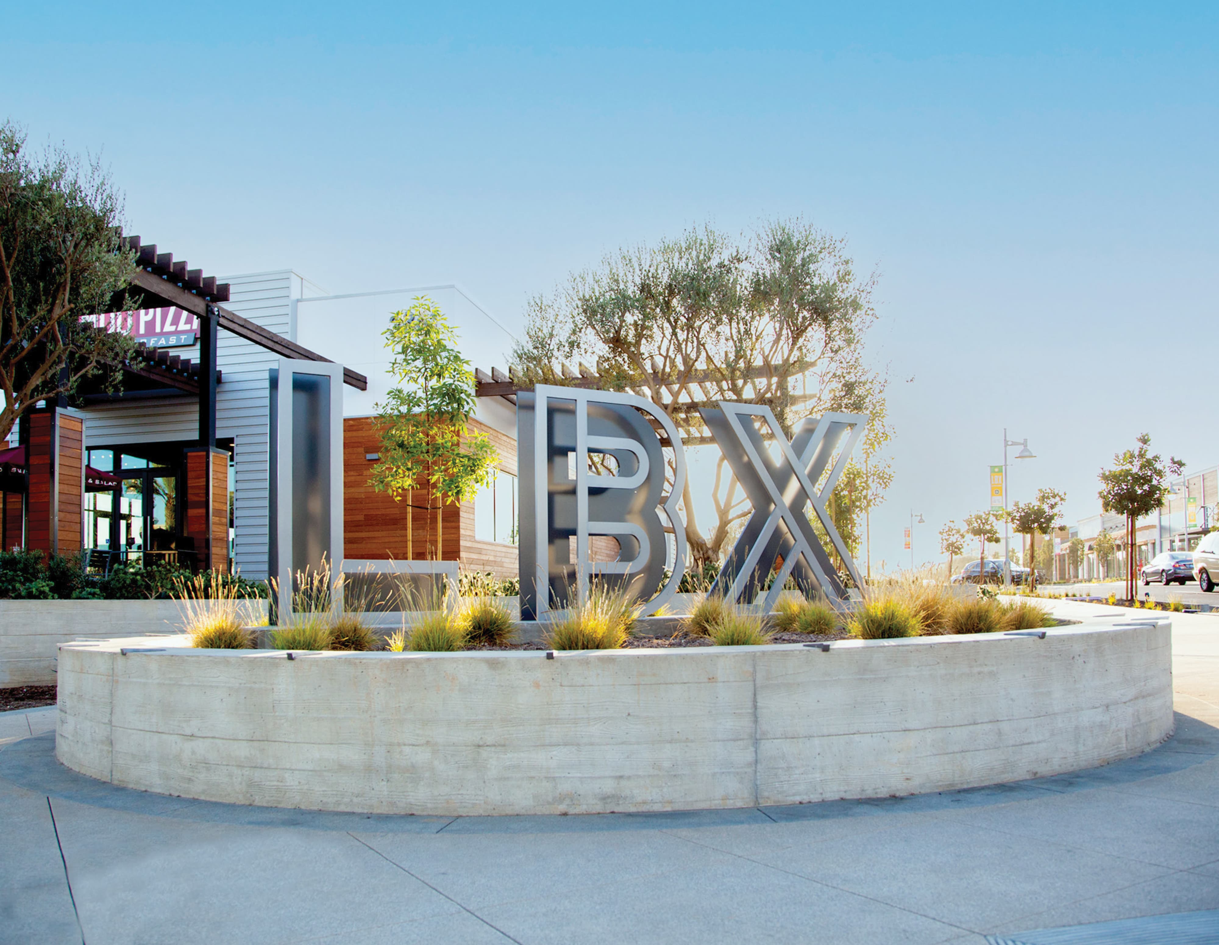 Close-up of a metal monument sign corner with perforated panels and an outlined 'LBX' logo, beside ornamental grasses and a red awning reading 'Fast Pizza & Salad' in the background.