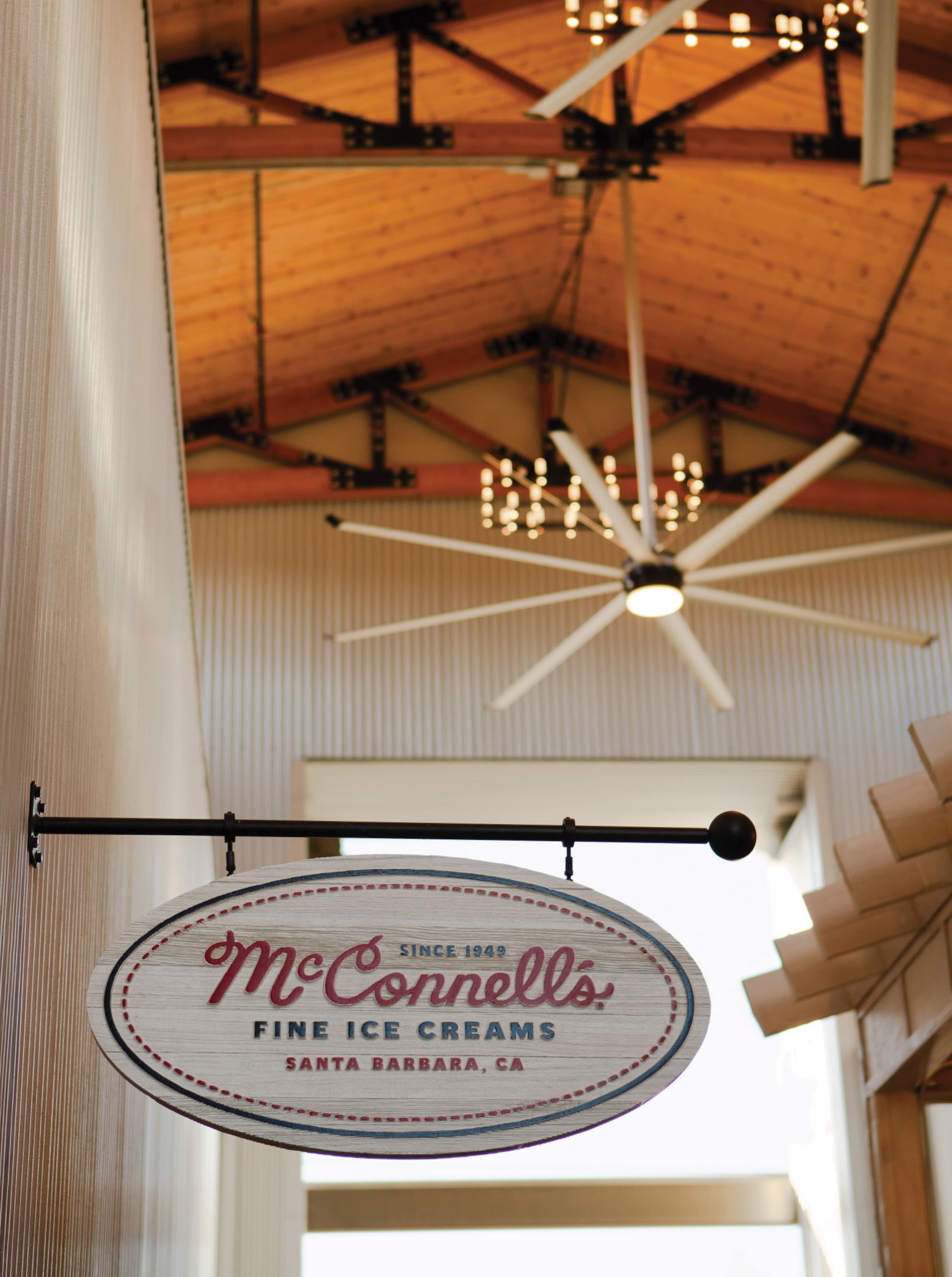 RSM Design tenant signage for McConnell's Fine Ice Creams at River Street Marketplace, featuring a close-up of a hanging oval blade sign with vintage script typography and distressed wood finish, suspended beneath an exposed wood beam ceiling, showcasing specialty graphics and branded environmental design for food and beverage and mixed-use retail environments.