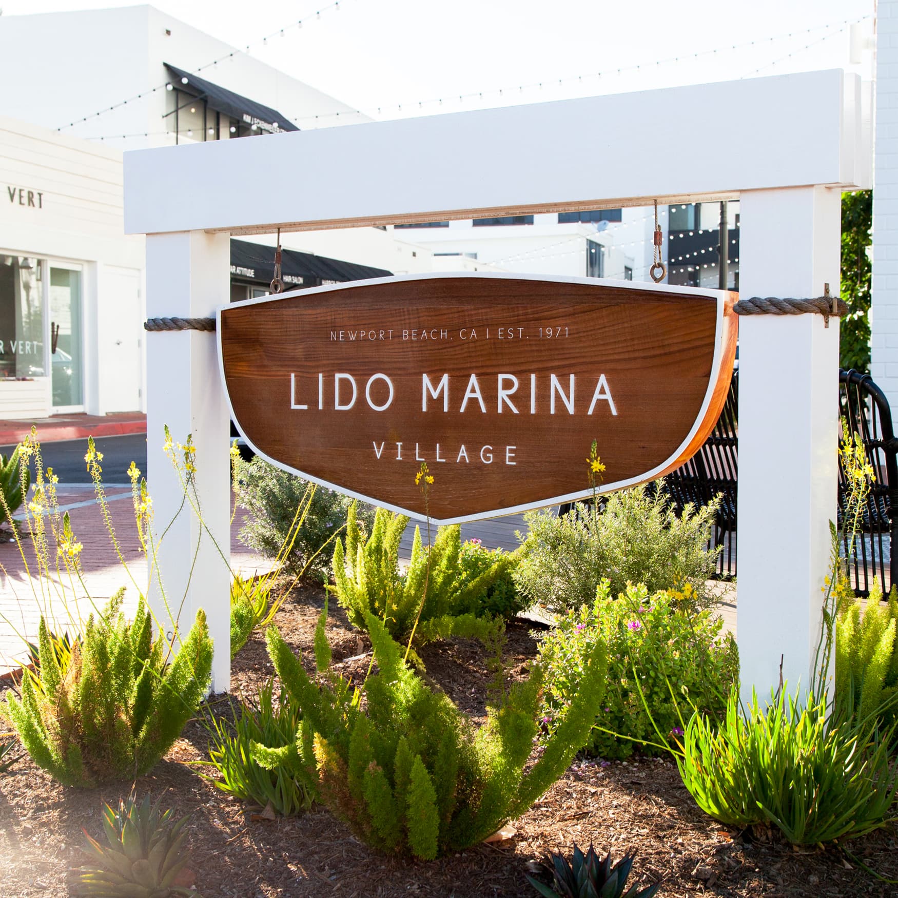 Lido Marina Village Sign in Newport Beach, CA

The image showcases the entrance sign for Lido Marina Village, located in Newport Beach, CA, established in 1971. The sign features a wooden panel with white lettering and is framed by a white structure with nautical rope details, evoking a coastal theme. The surrounding landscape includes neatly maintained plants and greenery, adding to the village's inviting and charming atmosphere. In the background, modern storefronts and string lights contribute to a lively and welcoming ambiance, reflecting the area's blend of contemporary style and seaside charm.