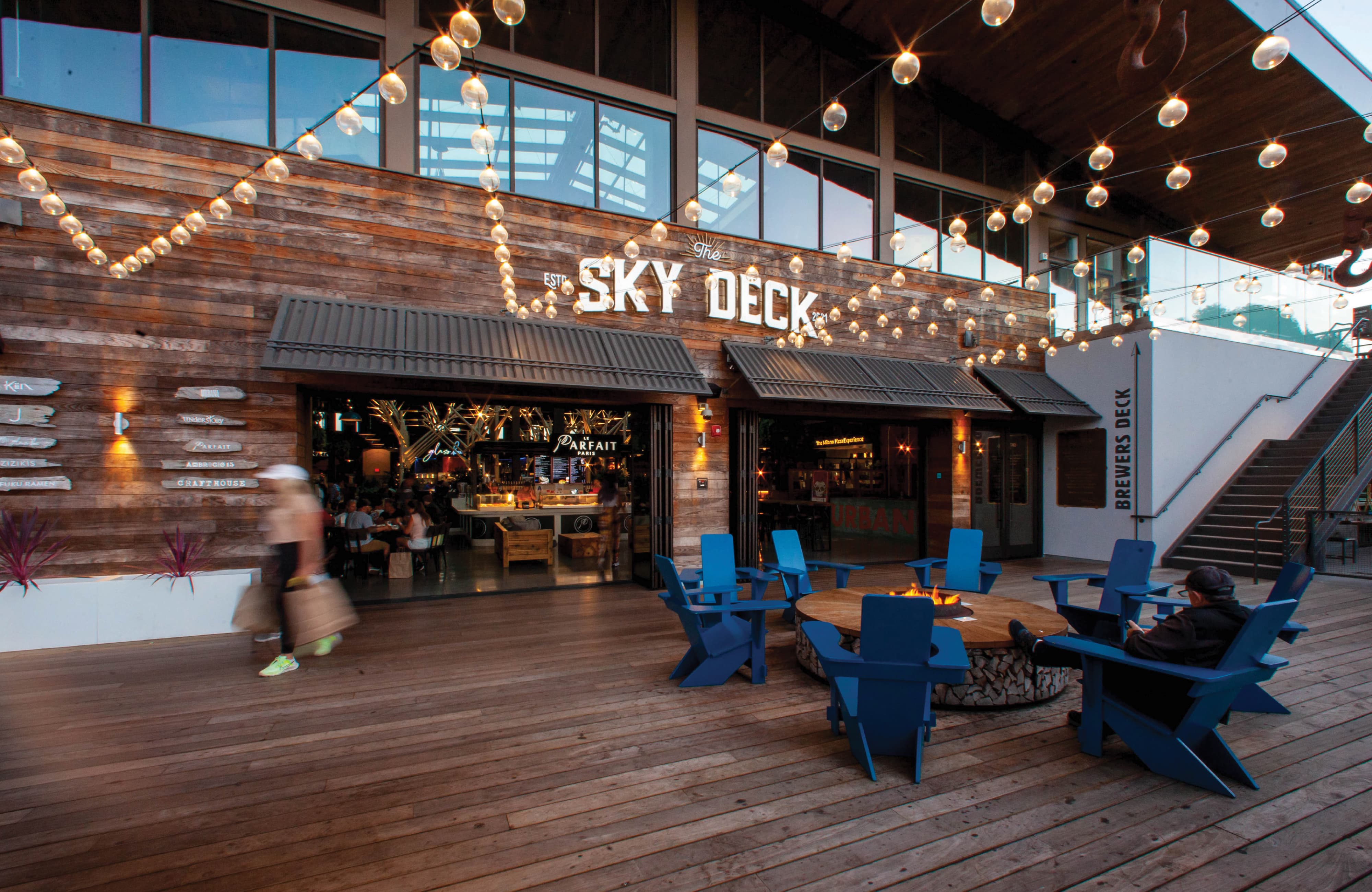 Environmental graphic design and branded placemaking at an outdoor hospitality and mixed-use destination, featuring large-scale building identification signage for The Sky Deck, integrated wayfinding, and experiential graphics. The open-air food hall environment includes custom lighting, tenant identity, and activation elements that enhance guest experience, support retail and entertainment uses, and strengthen sense of place within a coastal lifestyle destination.