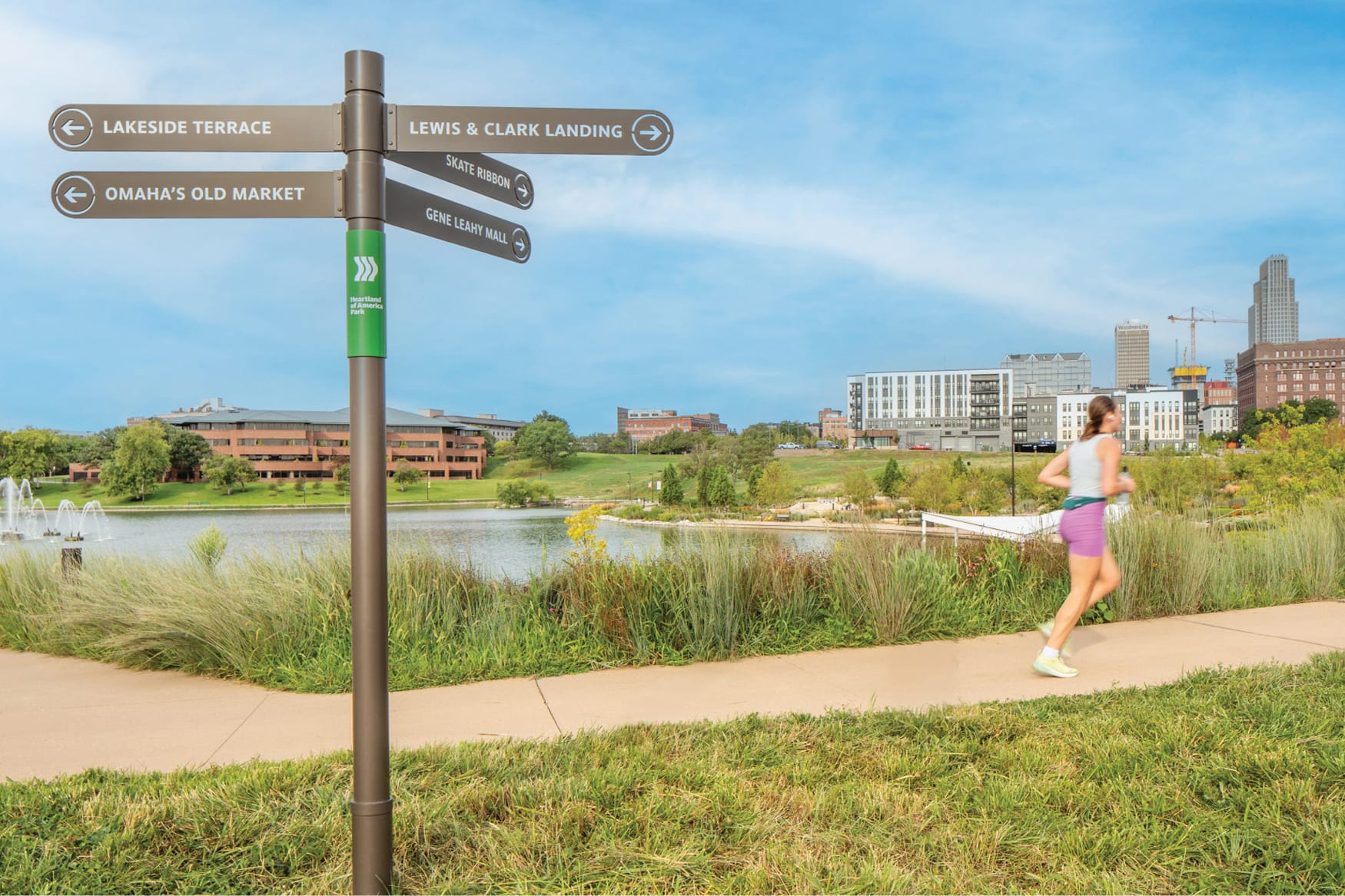 Heartland of America Park wayfinding sign in Omaha, Nebraska, directing visitors to Lakeside Terrace, Omaha's Old Market, Lewis and Clark Landing, Skate Ribbon, and Gene Leahy Mall, with a jogger, lakeside fountains, native grasses, and the downtown Omaha skyline in the background