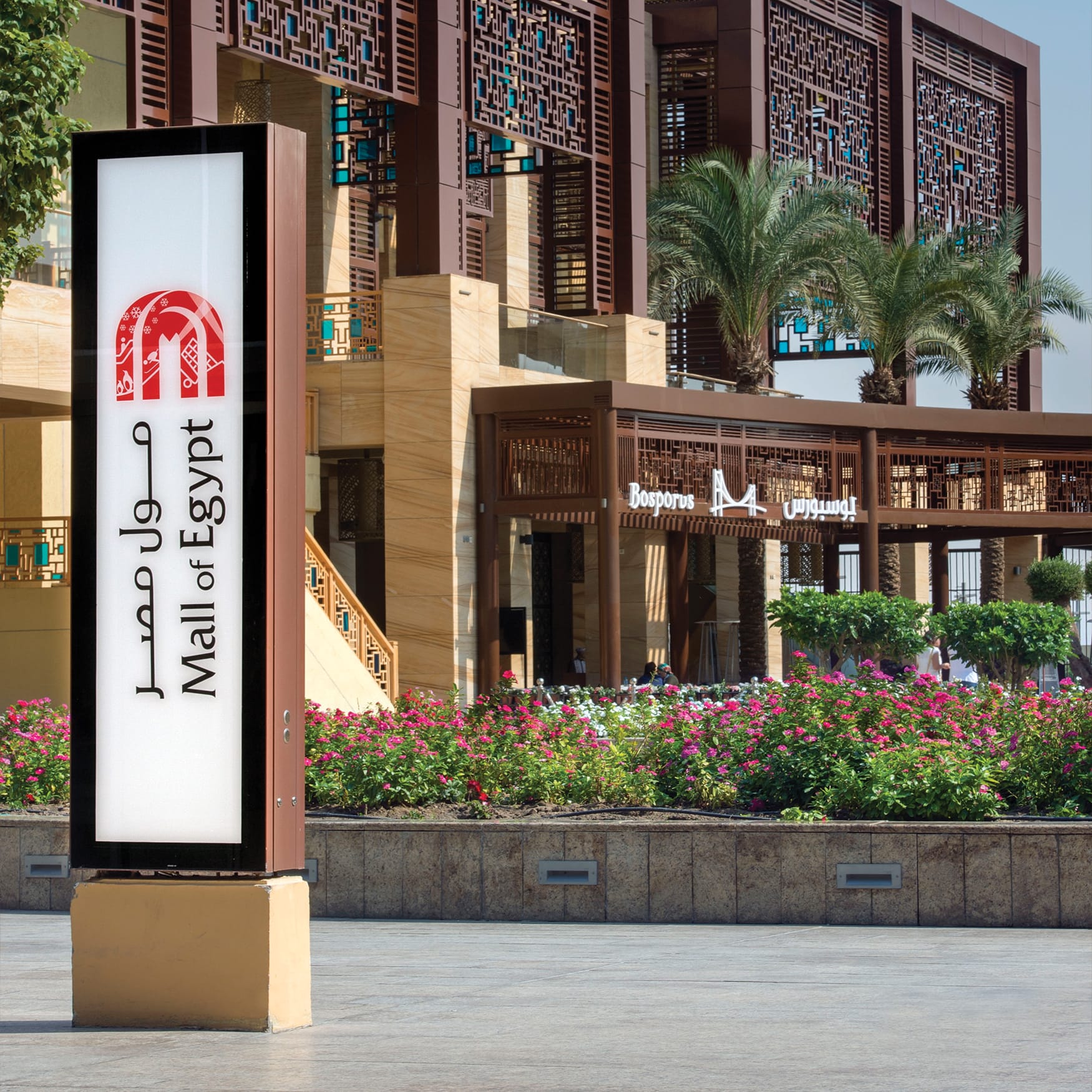 Exterior wayfinding sign for the Mall of Egypt featuring bilingual Arabic and English branding, illuminated signage, landscaped plaza, palm trees, and contemporary architectural details with geometric screens.