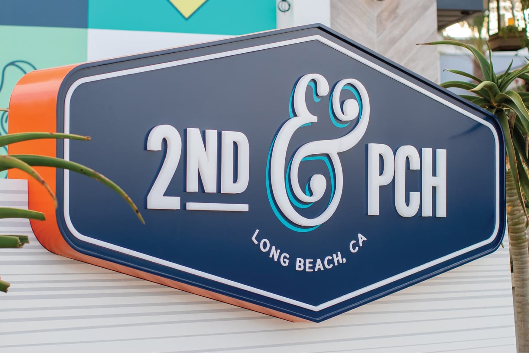Close-up of a dimensional monument sign for 2nd & PCH in Long Beach, California, featuring a navy and orange coffin-shaped cabinet with raised white lettering and a decorative teal ampersand — showcasing RSM Design's environmental graphic design, branding and visual identity, and placemaking for a coastal waterfront retail and mixed-use destination.