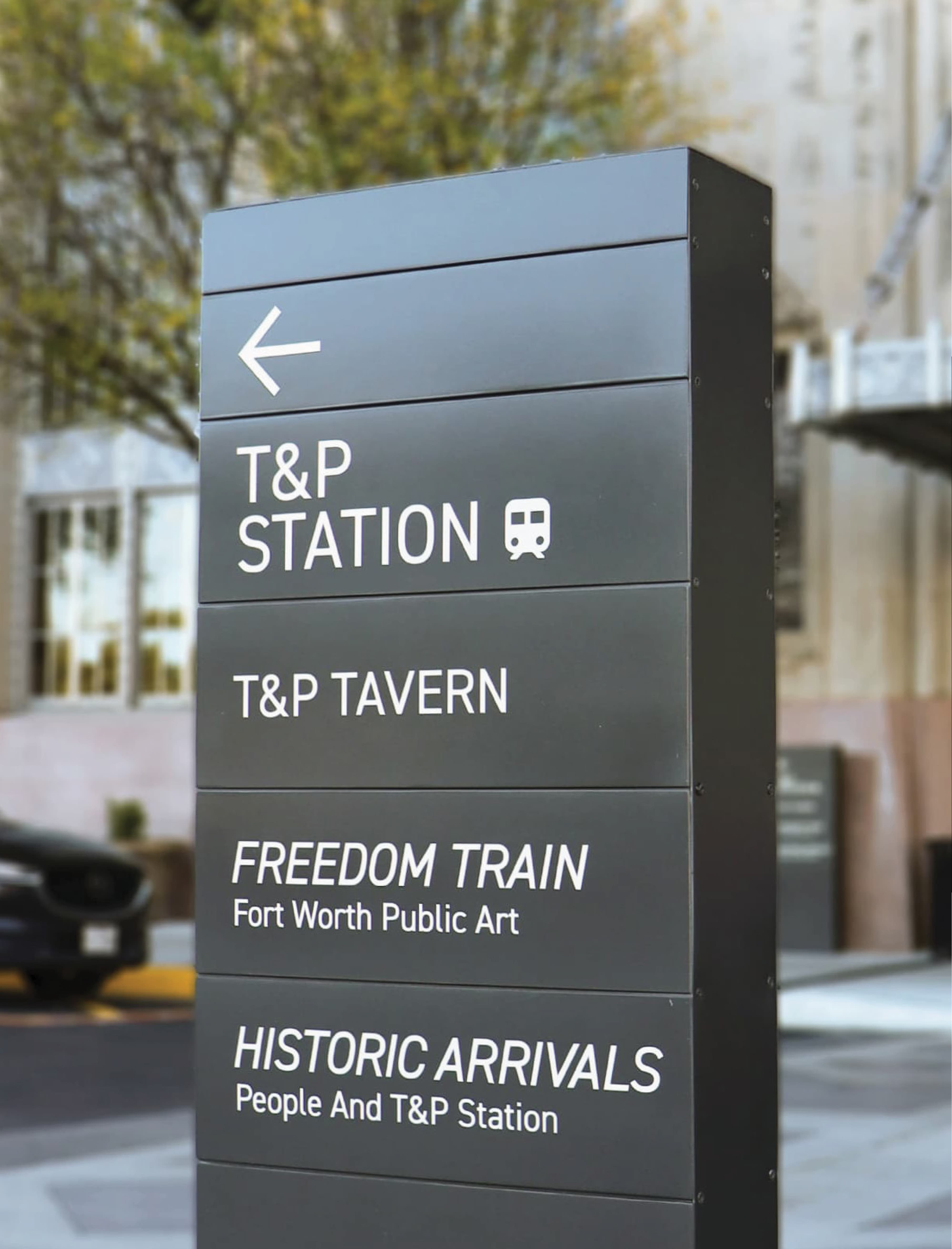 Dark charcoal pedestrian wayfinding totem directing visitors to T&P Station, T&P Tavern, Freedom Train Fort Worth Public Art, and Historic Arrivals on a downtown streetscape, showcasing environmental graphic design and signage for a mixed-use transit development by RSM Design.